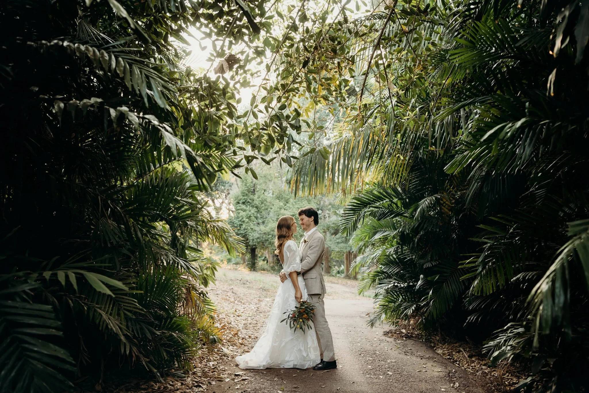 A bride and groom standing close together under a lush, green canopy in an outdoor setting, holding hands and looking into each other's eyes.