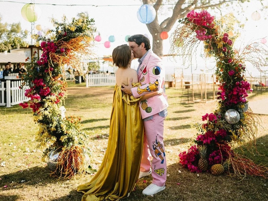 A bride and groom holding hands during an outdoor wedding ceremony with officiant and groomsmen, dressed formally, standing on grass beneath trees, with floral arrangements on white pedestals.