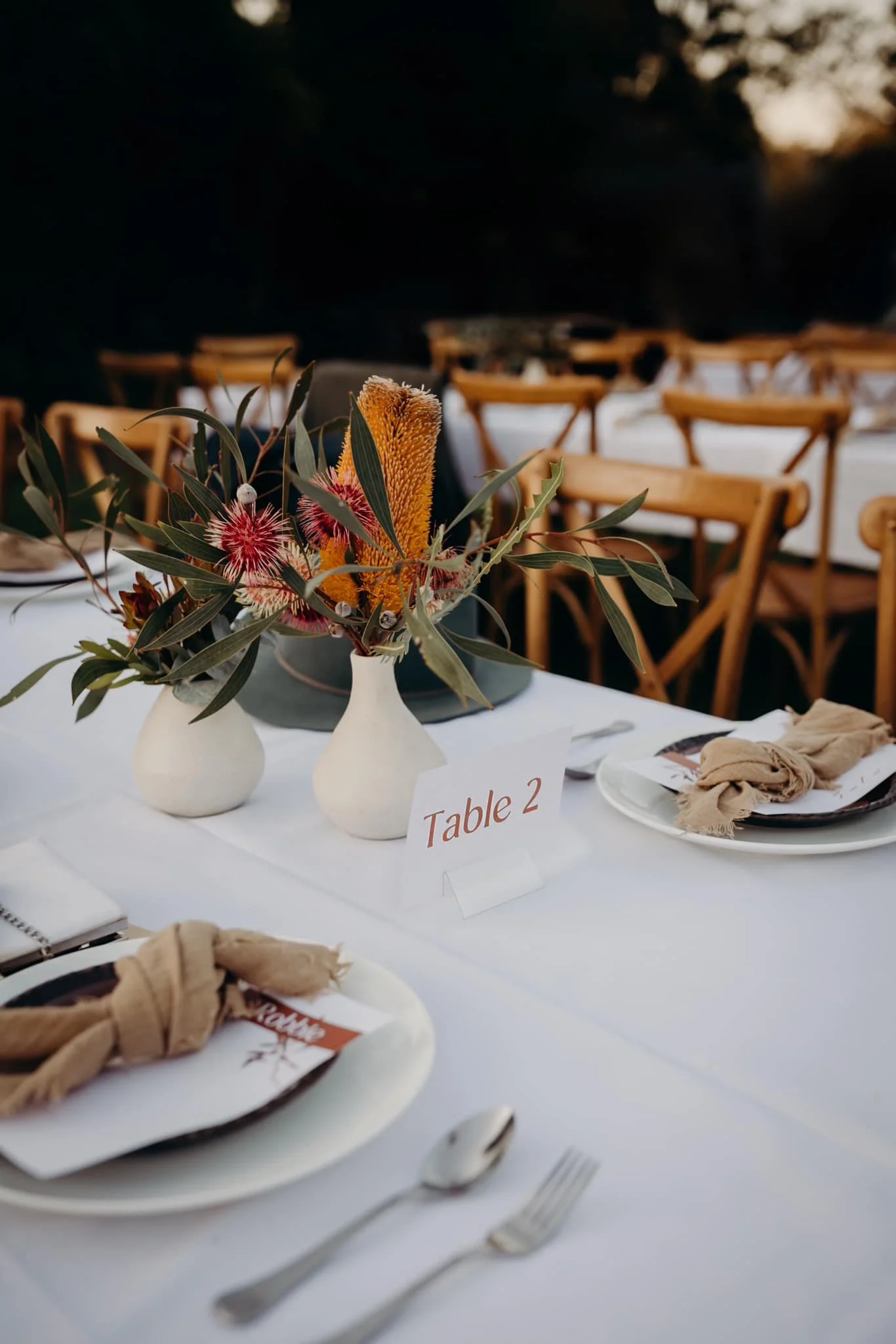 A floral centerpiece with various flowers and greenery on a table labeled 'Table 2' at a formal event. In the background, empty wooden chairs are arranged around other tables.