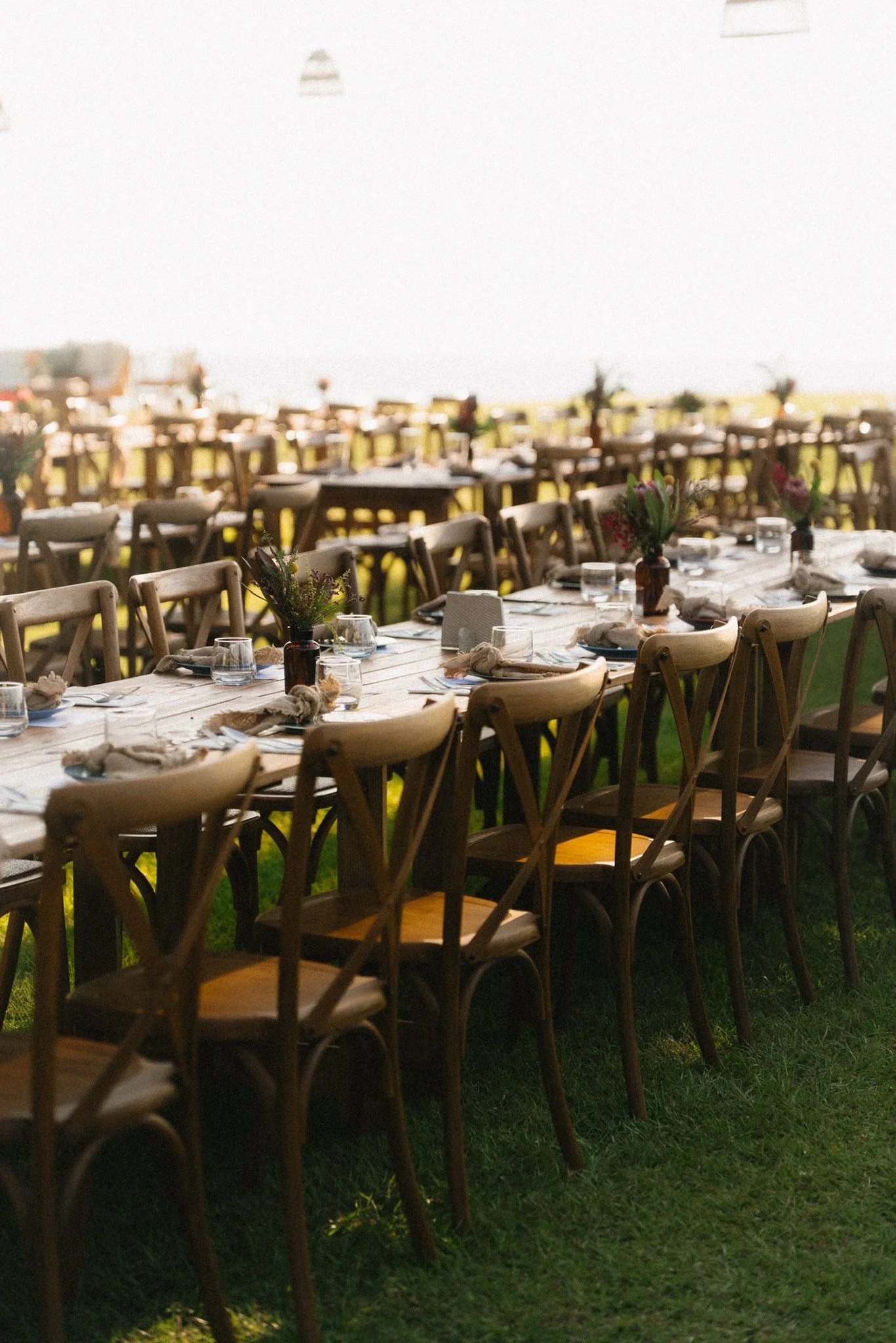 Long banquet table set with floral centerpieces, glasses, napkins, and plates in an outdoor setting during sunset.