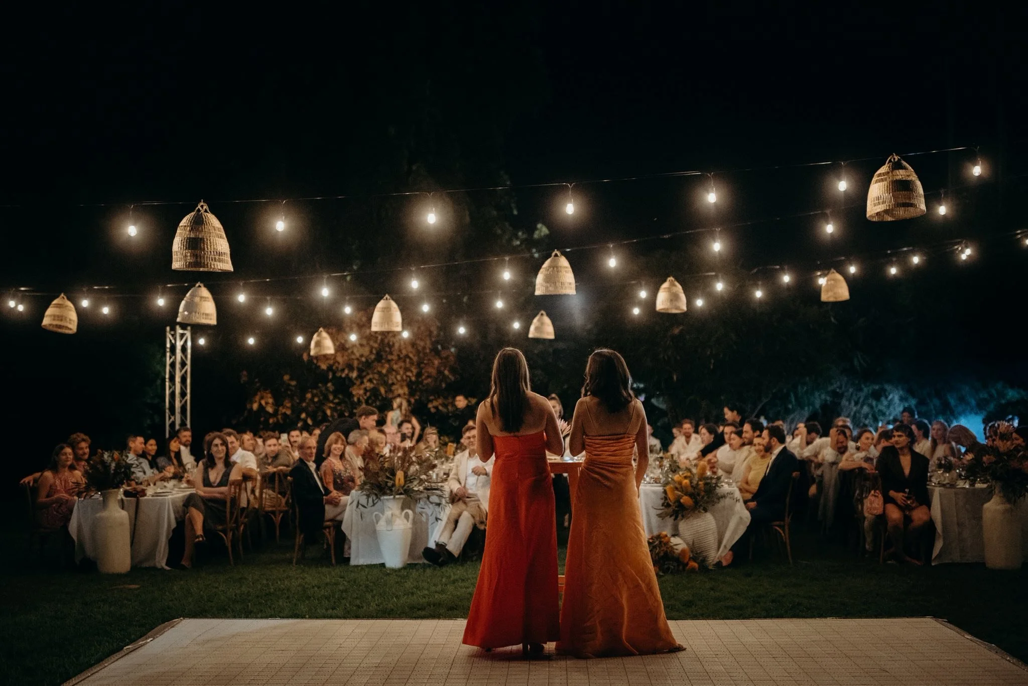 Nighttime outdoor wedding reception with hanging wicker lanterns, decorated tables, and numerous guests seated, with two women in dresses speaking at a podium.