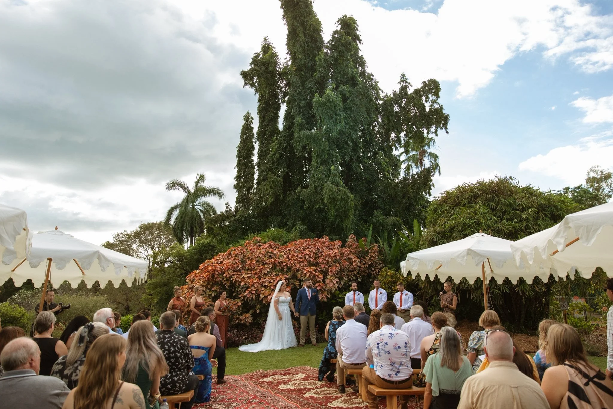 An outdoor wedding ceremony with a bride and groom standing in front of a large tree, surrounded by wedding guests. The sky is partly cloudy, and white umbrellas are providing shade for the guests.
