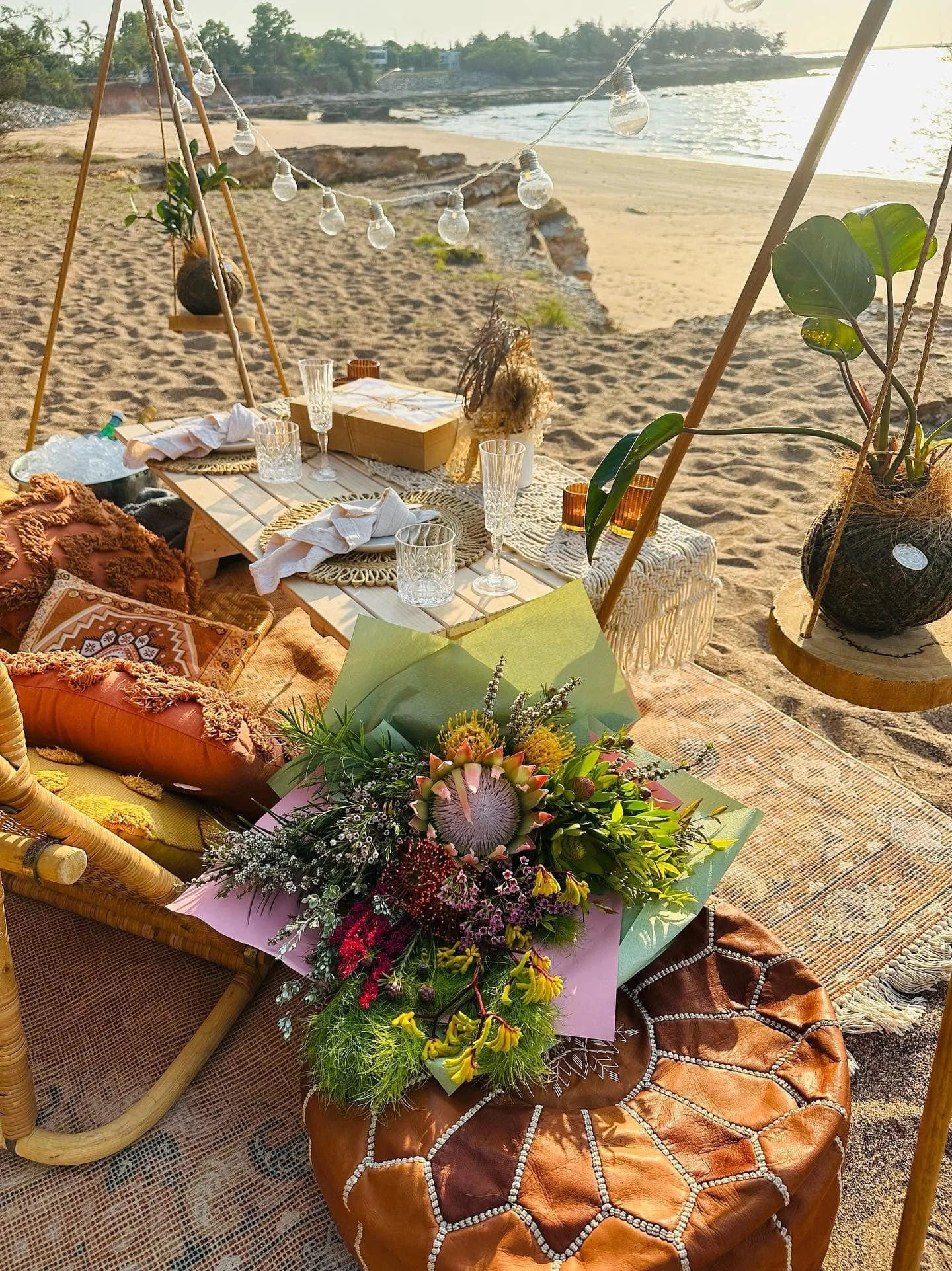 A beachside dining setup with a low wooden table, glassware, and a bouquet of flowers. There are cushions and throws on the sandy ground, and string lights are hung between wooden poles. The ocean and sandy beach are visible in the background.