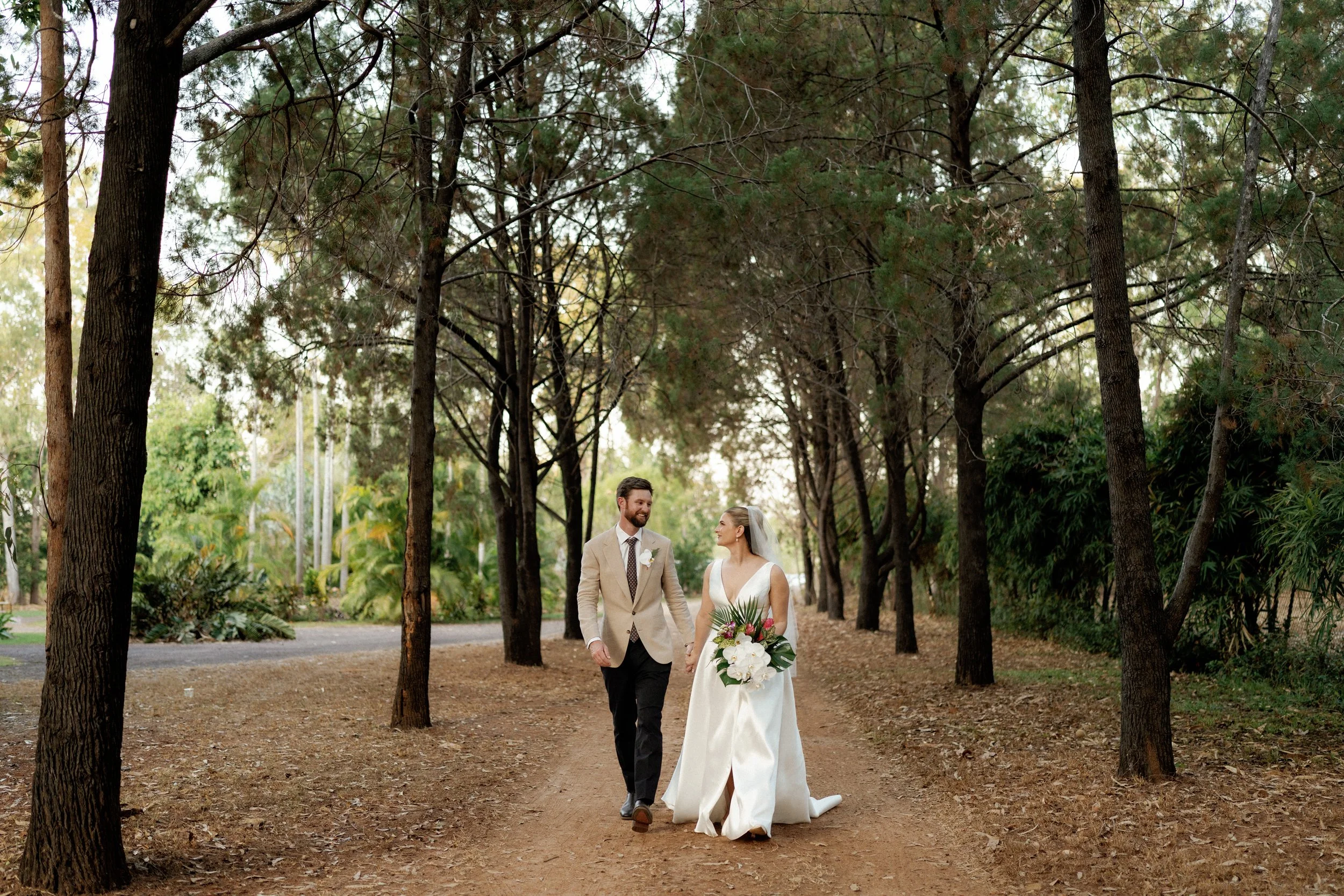 An outdoor wedding ceremony with a bride and groom standing in front of a large tree, surrounded by wedding guests. The sky is partly cloudy, and white umbrellas are providing shade for the guests.