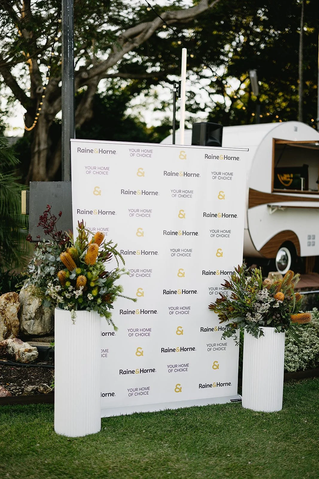 A promotional backdrop with logos for Raine & Horne and text 'Your Home of Choice,' accompanied by two large flower arrangements in white vases on a grassy area. In the background, there is a small white and wooden travel trailer.