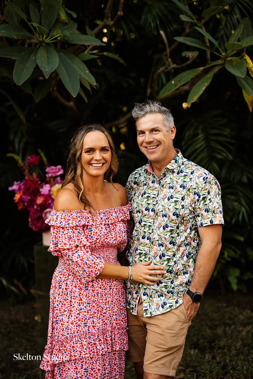 A smiling couple standing close together outdoors, surrounded by lush green foliage and pink flowers, with the woman wearing a pink off-shoulder dress and the man in a colorful shirt and khaki shorts.