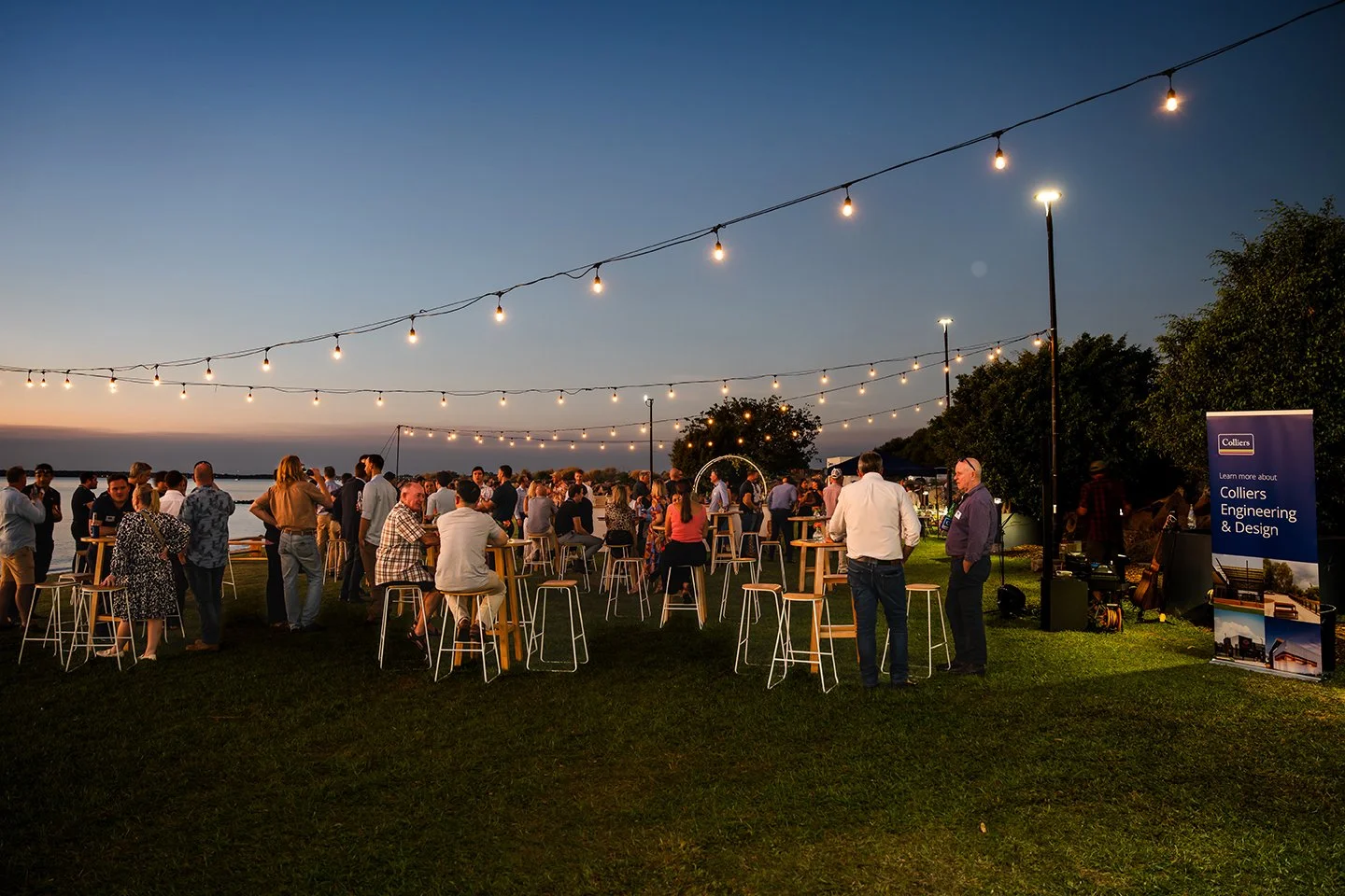 Outdoor evening social gathering with people standing and sitting around high tables, string lights overhead, near water, with a sign for Colliers Engineering & Design.
