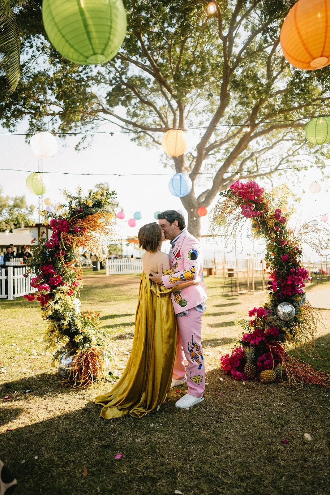 A couple kissing under a decorated arch at an outdoor celebration with colorful lanterns hanging from trees.