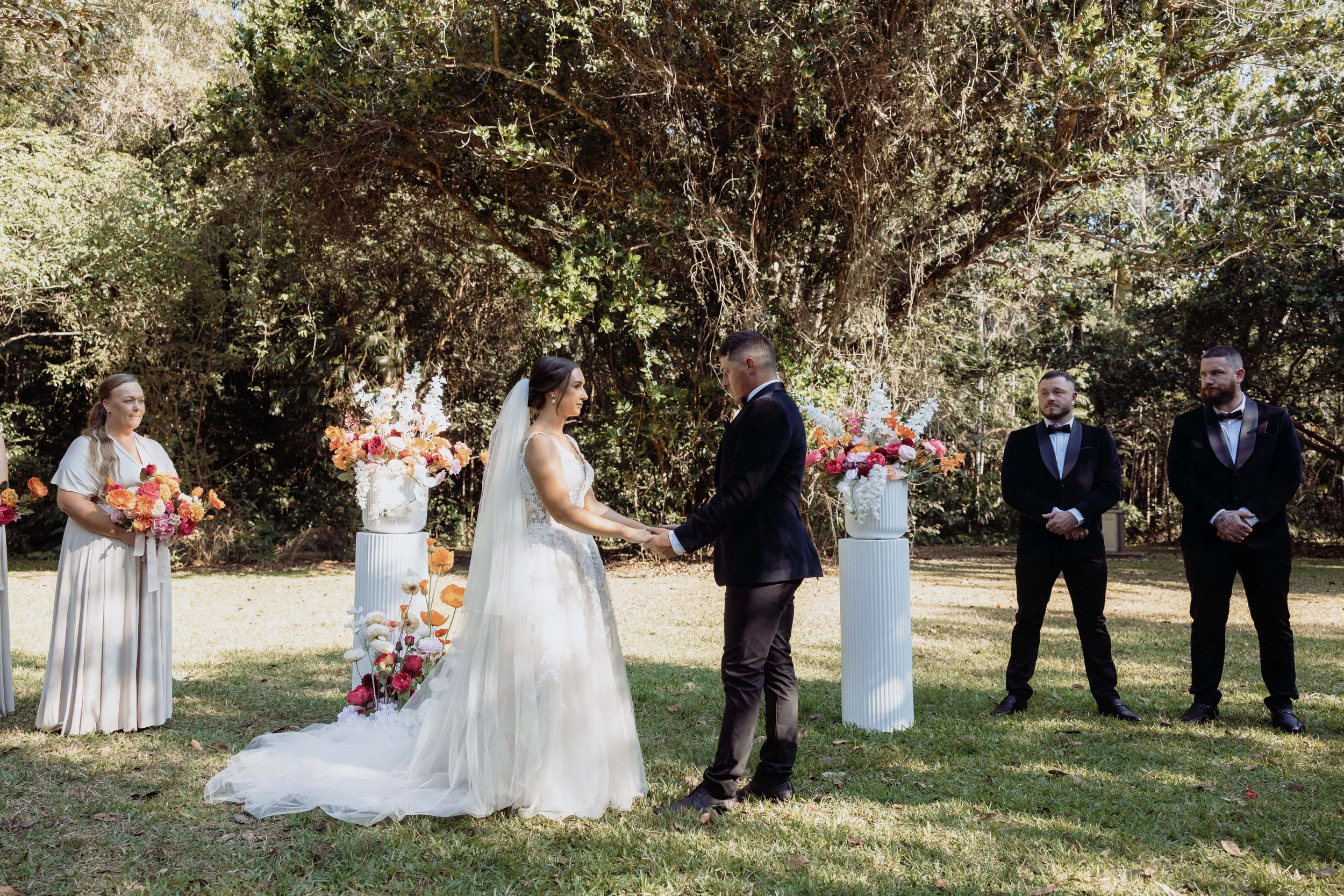 A bride and groom holding hands during an outdoor wedding ceremony with officiant and groomsmen, dressed formally, standing on grass beneath trees, with floral arrangements on white pedestals.