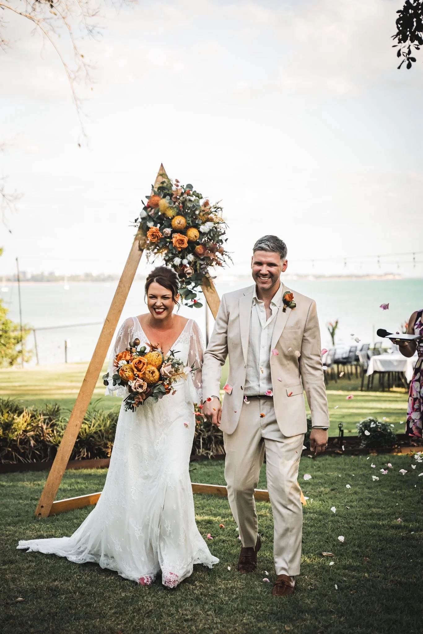 A smiling bride in a white lace wedding dress holding a bouquet of orange, yellow, and white flowers, walking hand-in-hand with a groom in a beige suit near a decorated wedding arch with flowers, outdoors by a body of water with a bridge in the background.