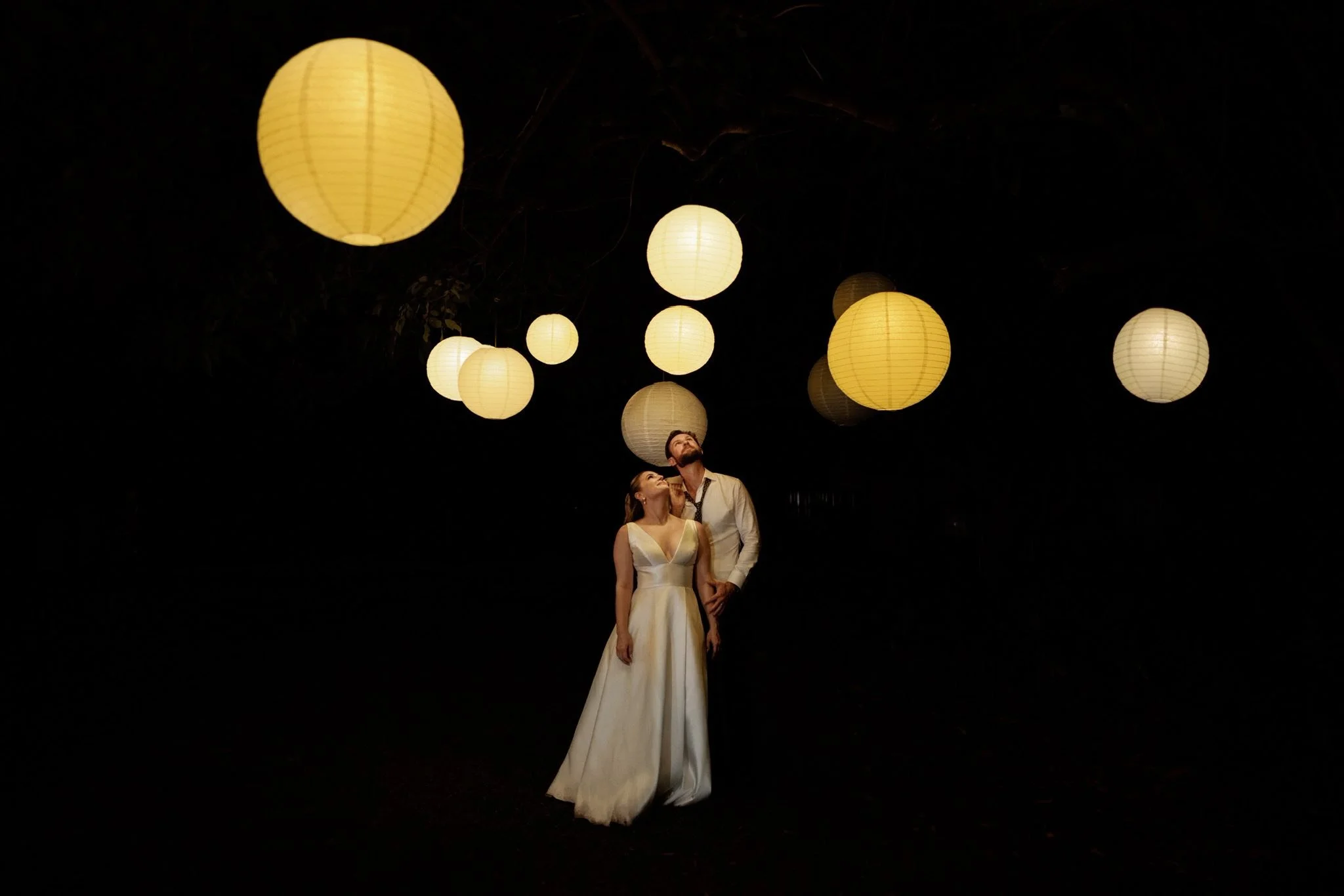 A couple dressed in formal attire stands underneath hanging paper lanterns at night, illuminated against a dark background.