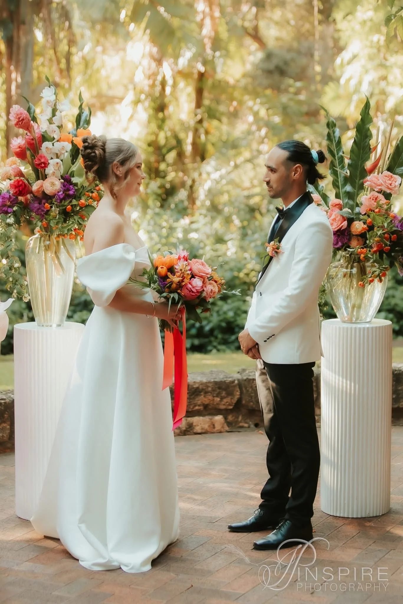 A bride and groom standing outdoors during a wedding ceremony, surrounded by flowers in vases, with trees in the background.