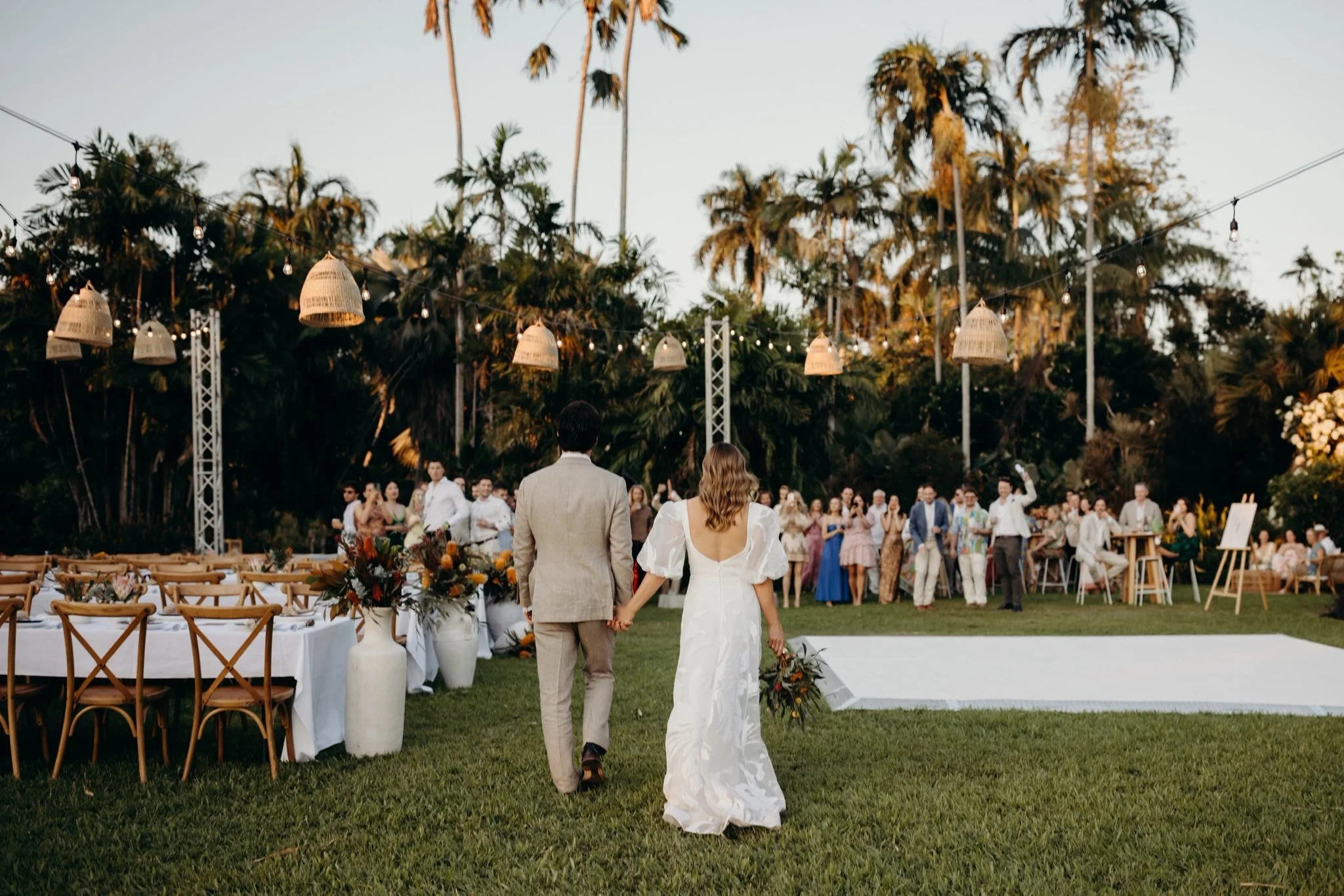 A bride and groom walking hand in hand at their outdoor wedding reception, with guests in the background, set amidst tall trees and hanging lanterns during sunset.