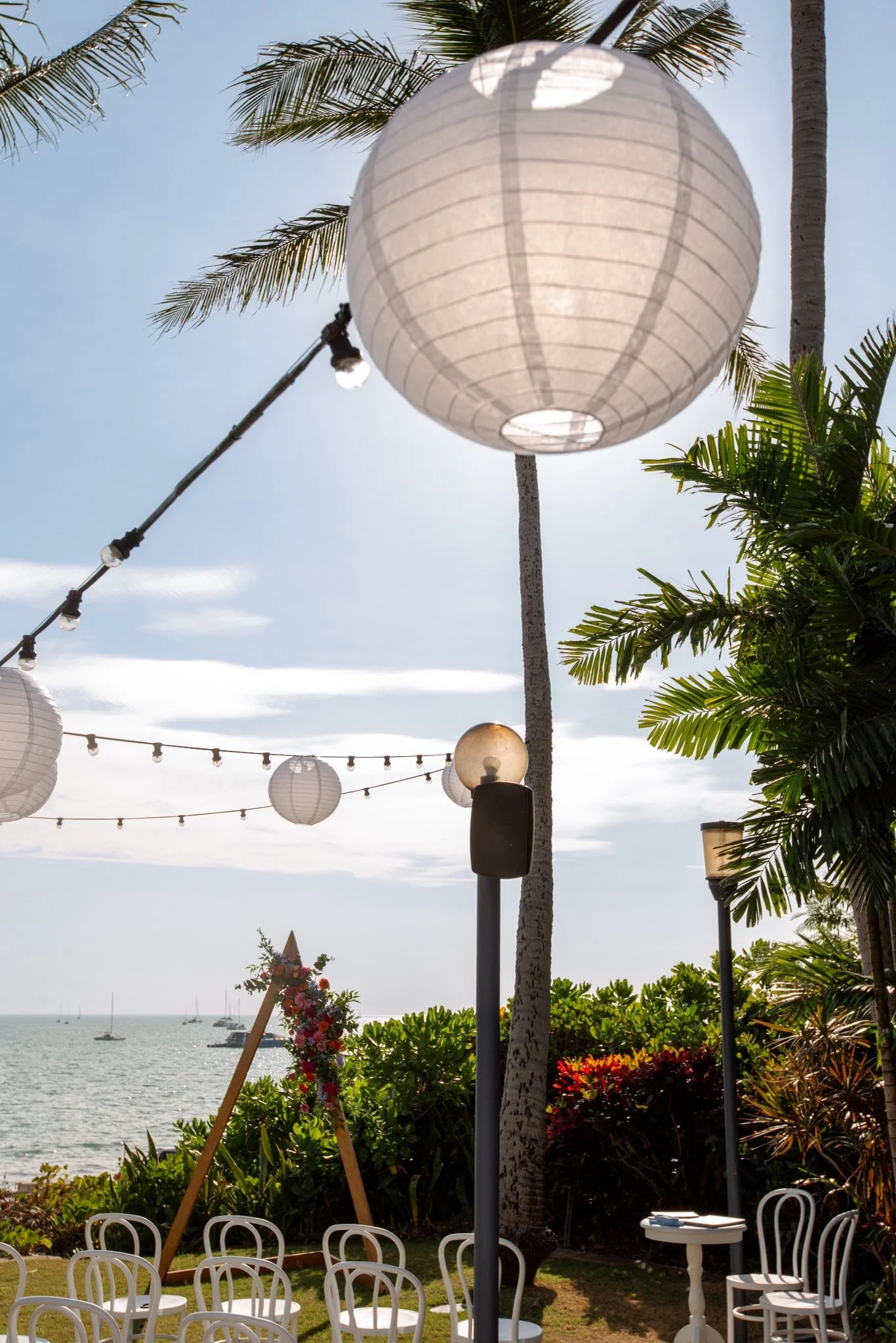 Outdoor event setup near the water with white chairs, paper lanterns hanging, palm trees, and sailboats in the distance.
