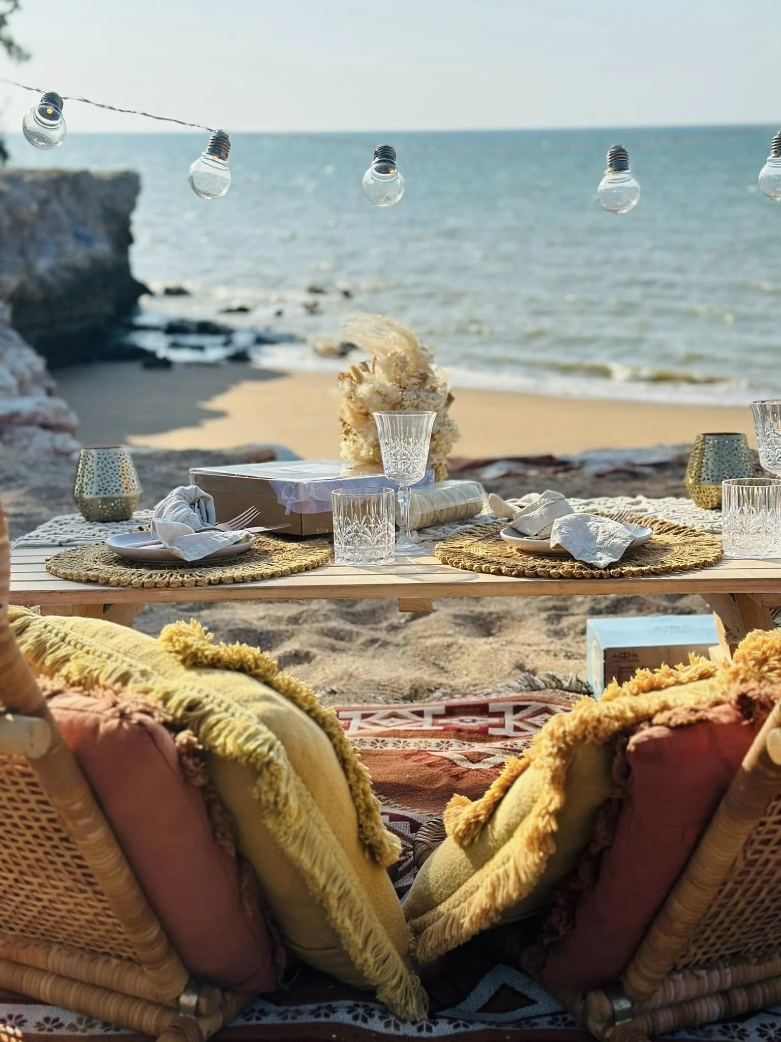 Outdoor beach scene with a low wooden table set for a meal, decorated with glasses, plates, and napkins, with a string of clear light bulbs hanging above, and a sandy shoreline with rocks and the ocean in the background.