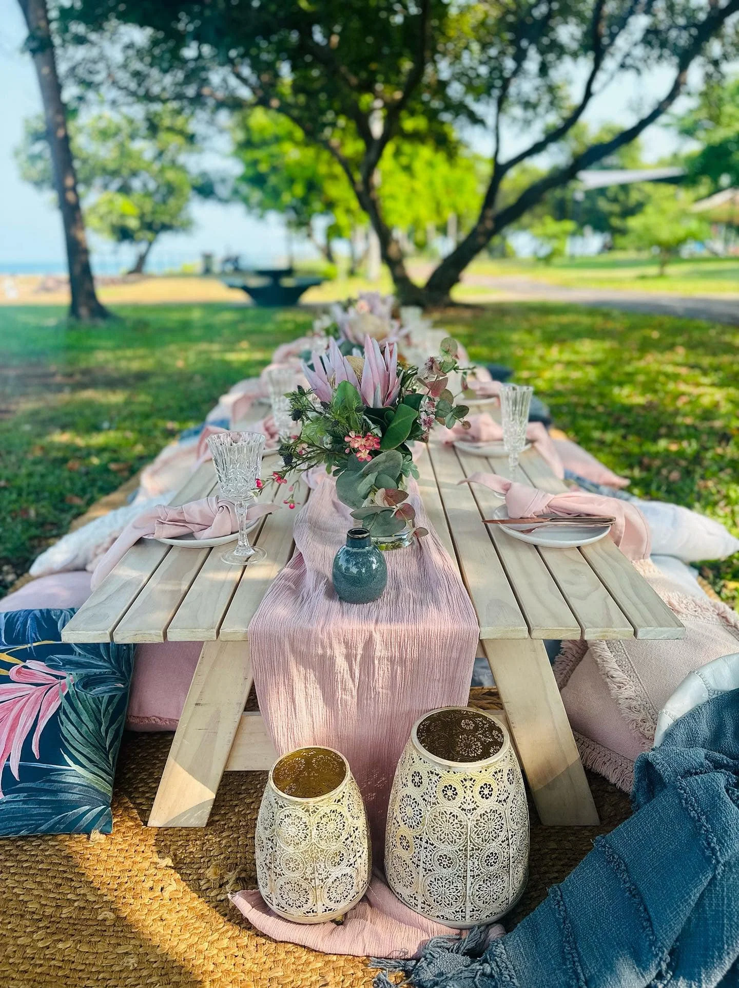 A picnic table decorated with pink and white flowers, surrounded by colorful pillows, lanterns, and cloths, set outdoors in a lush green park with trees and a pathway.
