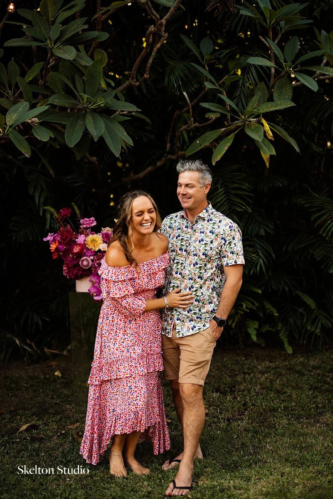 Smiling woman in pink patterned dress and man in floral shirt and beige shorts standing together outdoors near large leafy plants with a bouquet of pink flowers in the background.