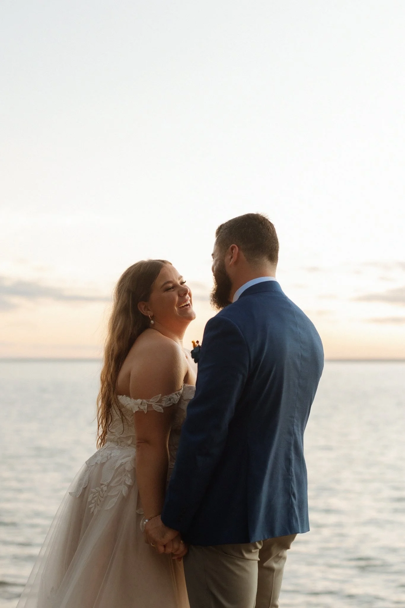 A bride and groom standing close together, holding hands and smiling at each other, with a body of water and a cloudy sky in the background during sunset.