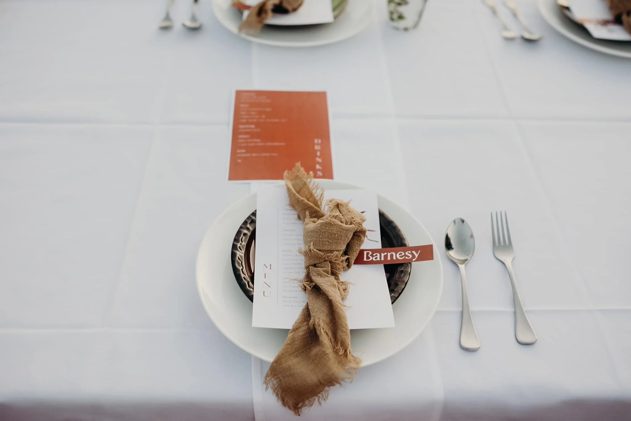 A table setting with a white plate, a beige cloth napkin tied with a string, and a restaurant menu labeled 'Barnesy.' The table has a white surface with utensils including a knife, fork, and spoon neatly placed beside the plate. In the background, there are additional plates and utensils.