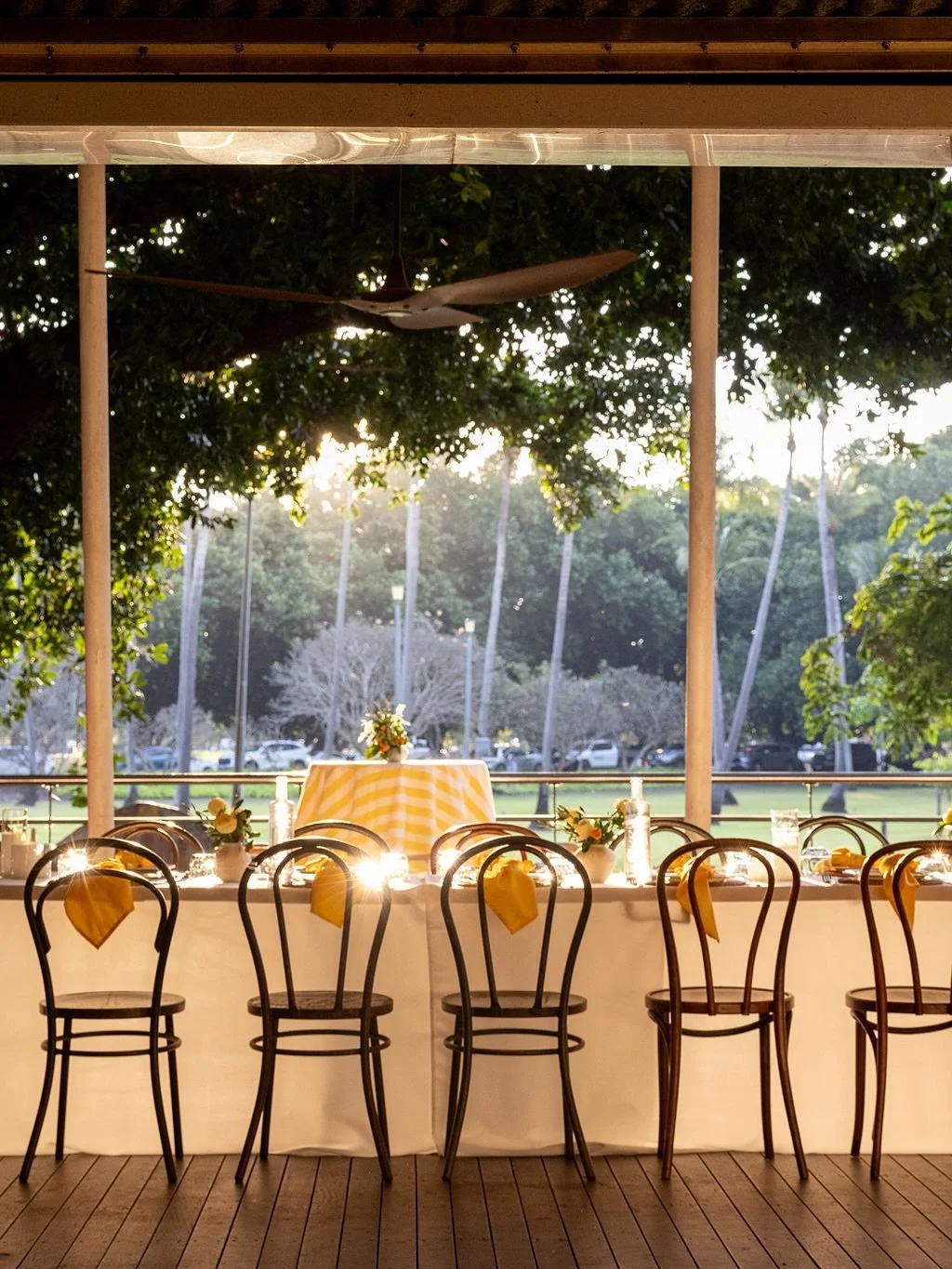Empty decorated dining table with chairs, white tablecloth, yellow napkins, flowers, and candles at an outdoor venue during sunset.