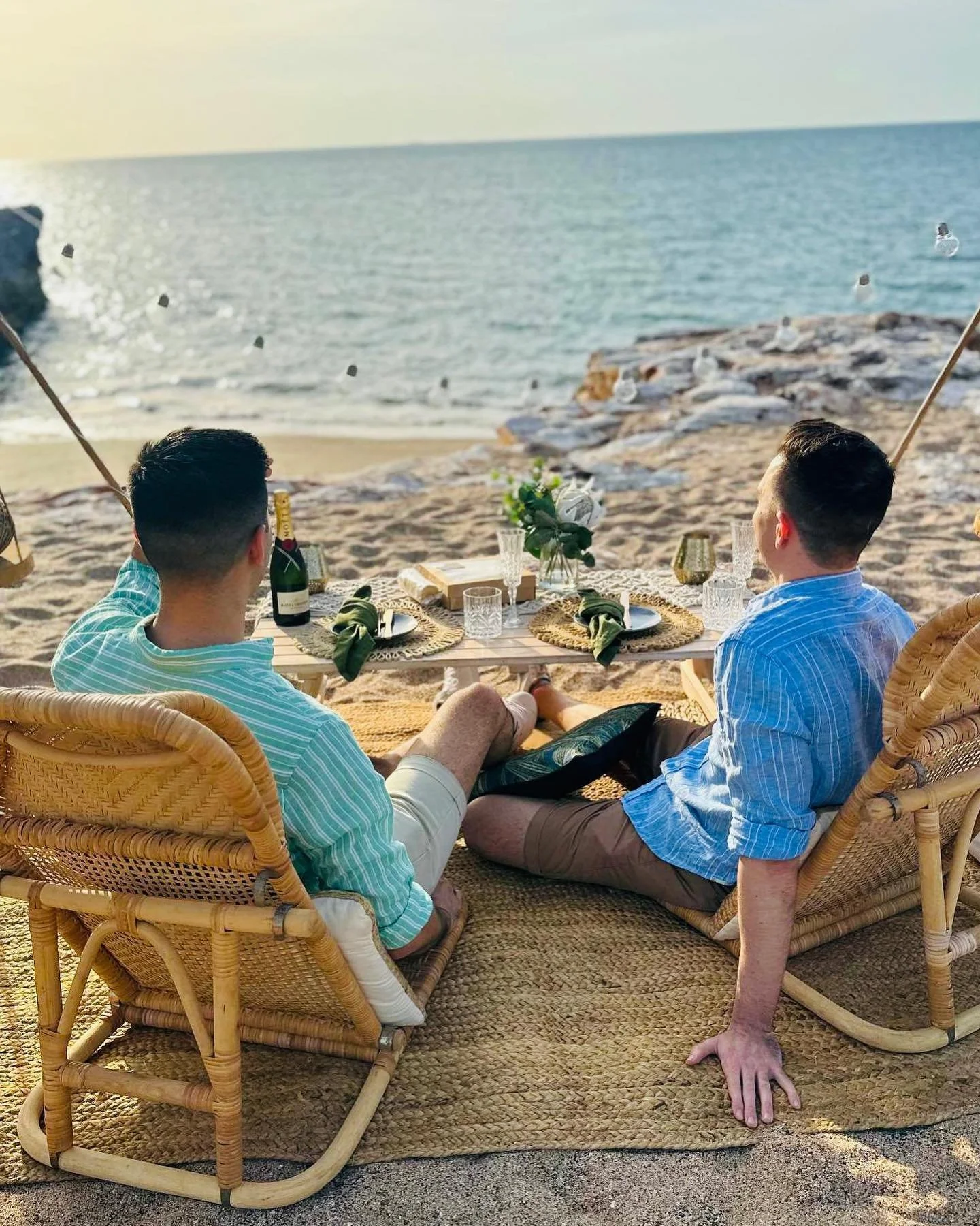 Two men sitting on a beach under a canopy, facing the ocean with rocks, a distant pier, and seagulls flying overhead, enjoying drinks and a meal.