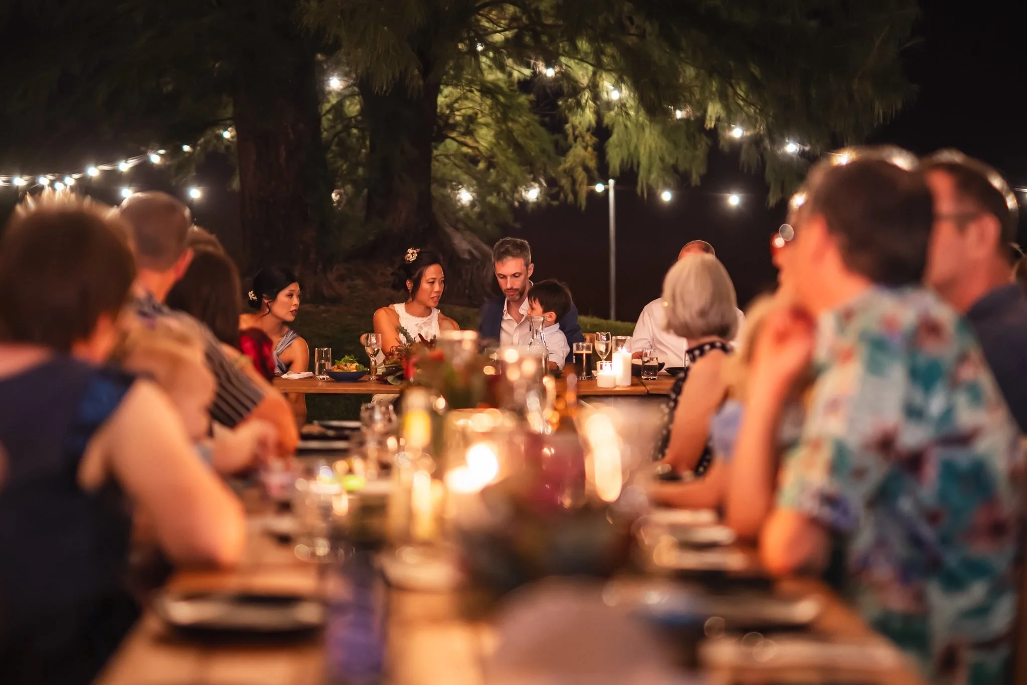 People dining outdoors at night, sitting around a long table illuminated by candles, with string lights and large trees in the background.