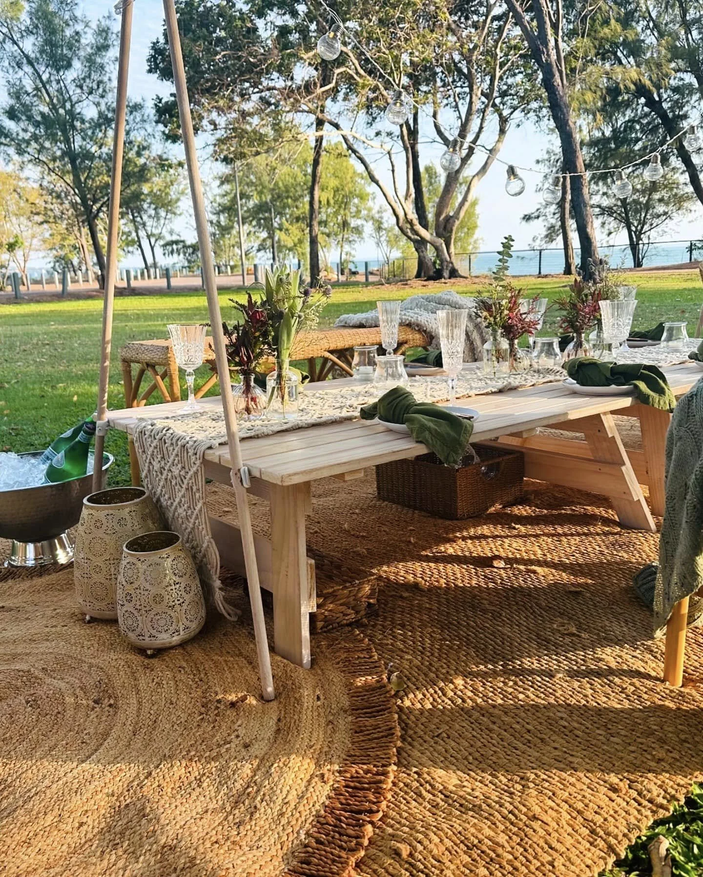 Outdoor dining setup on a wooden table with glassware, flowers, and cloth napkins, under string lights among trees on a grassy lawn with ocean in the background.