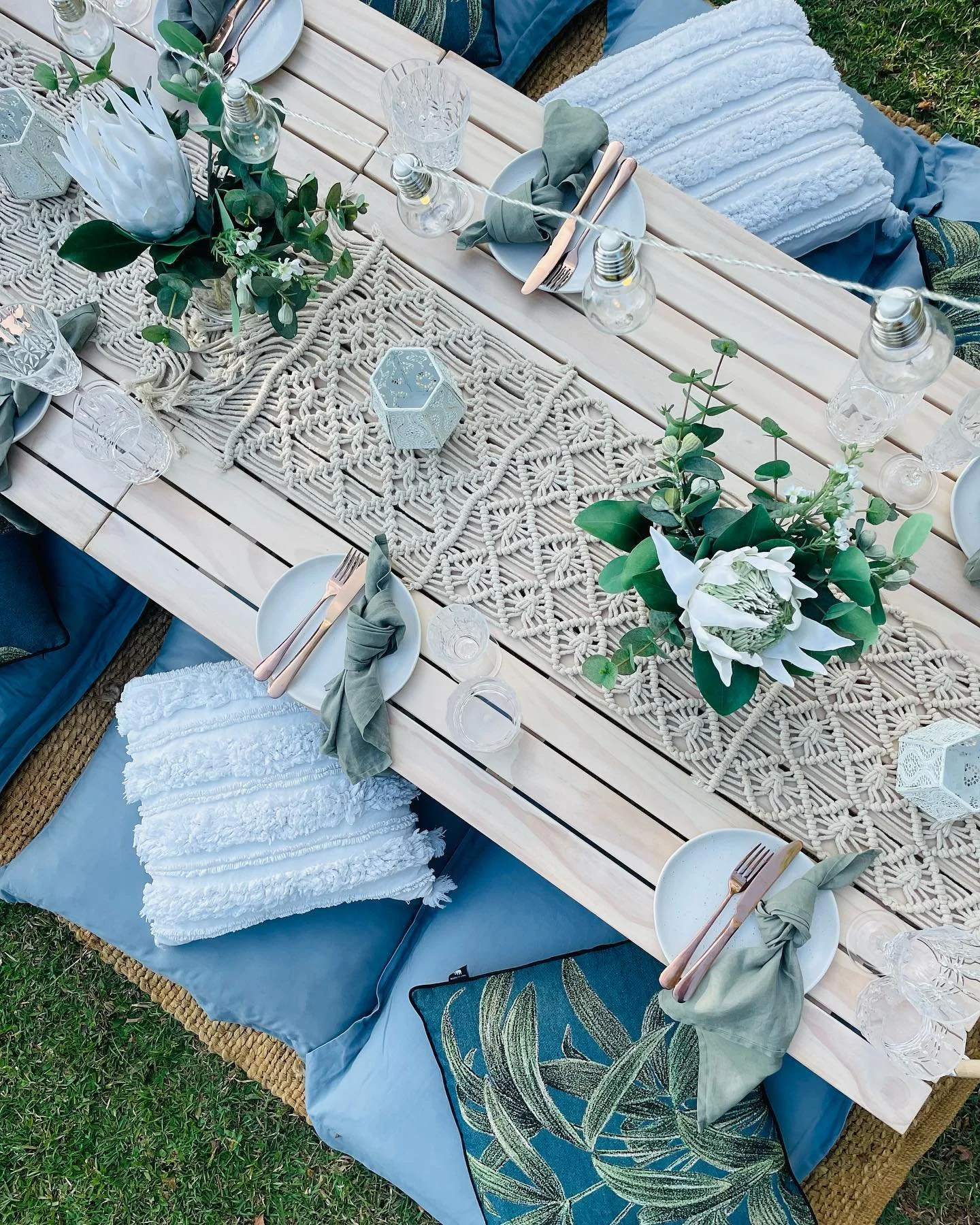 Outdoor dining table decorated with a floral centerpiece, string lights, white plates, cutlery wrapped with fabric napkins, glassware, and a woven table runner, set on a grassy area with pillows and textured throws.