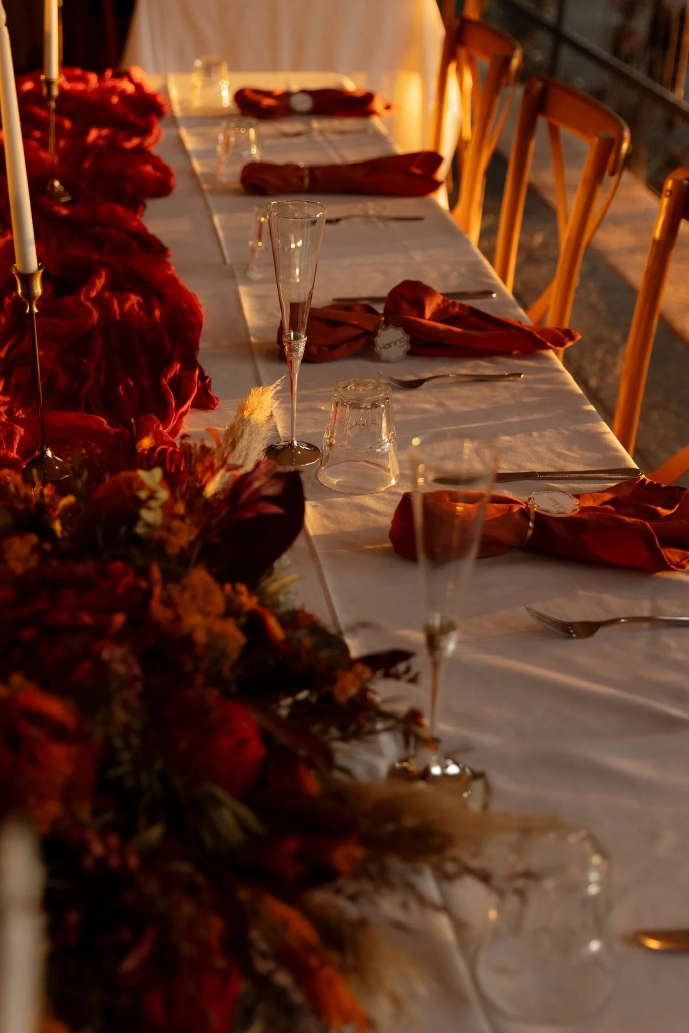 A long dining table set for a formal event with gold chairs, orange napkins, glassware, and a floral centerpiece, illuminated by warm lighting.