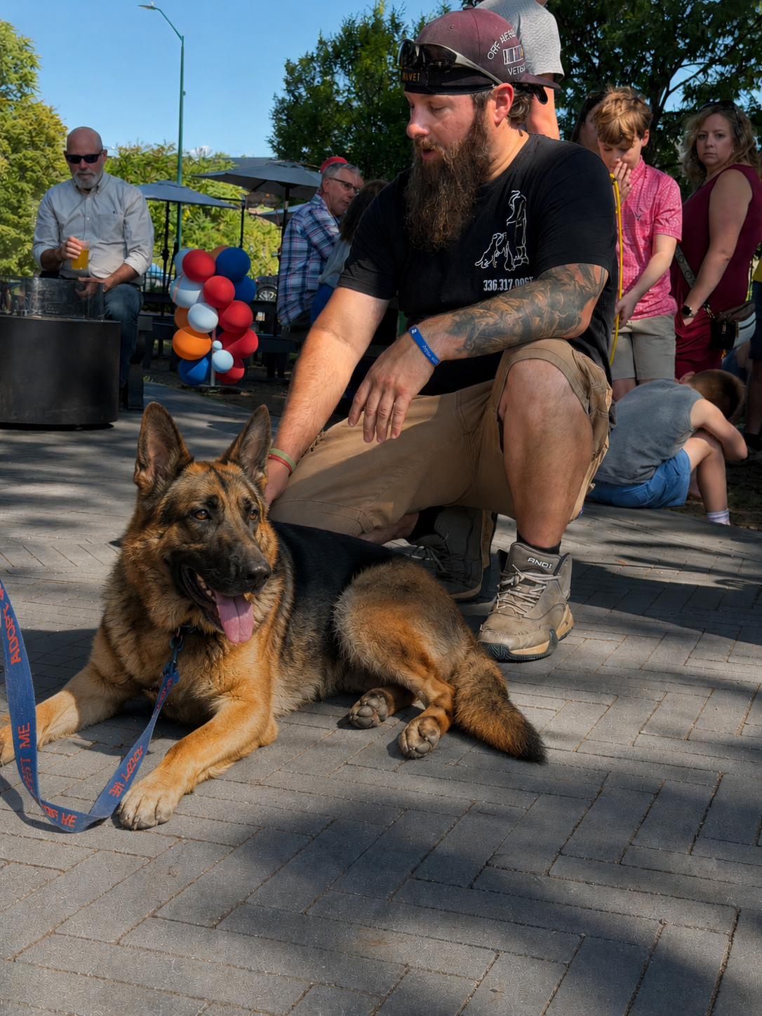 A man with a beard, wearing a black shirt, tan shorts, and hiking boots, is kneeling on the ground petting a German Shepherd dog lying on its side, in a crowded outdoor gathering with colorful balloons, people standing and sitting, some holding drinks, and a clear blue sky.