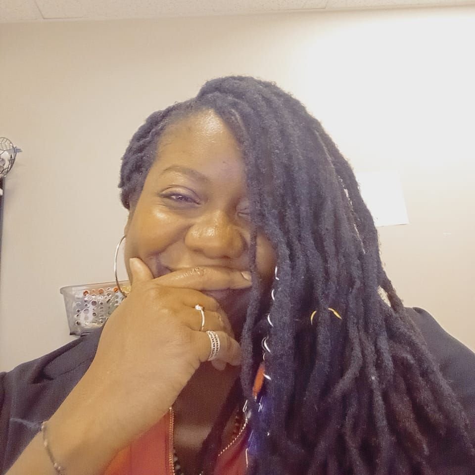 A woman with long, twisted dreadlocks, wearing hoop earrings, a ring, and a black jacket, smiling with her hand resting on her chin.