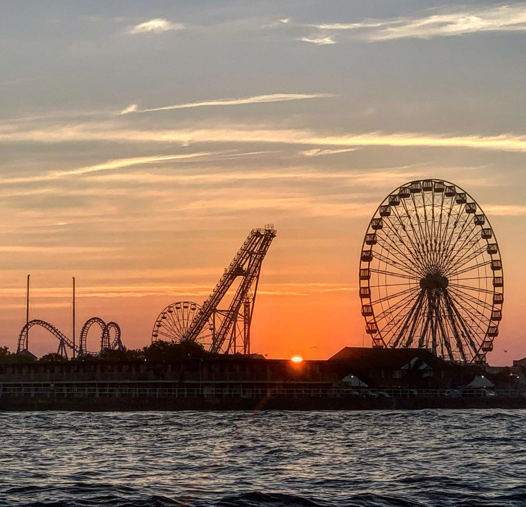 The image shows a Ferris wheel and amusement park rides silhouetted against a sunset sky with colorful clouds over a body of water.