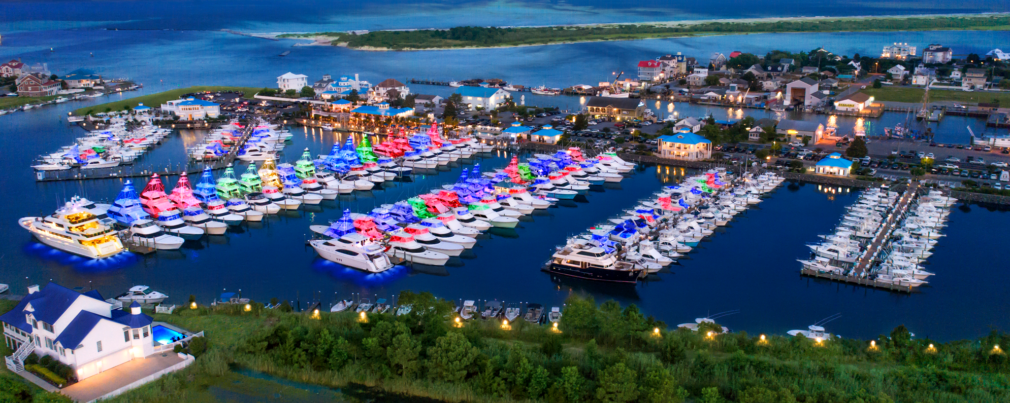 Aerial view of a marina at dusk decorated with colorful lights on boats, with a waterfront town and waterway in the background.