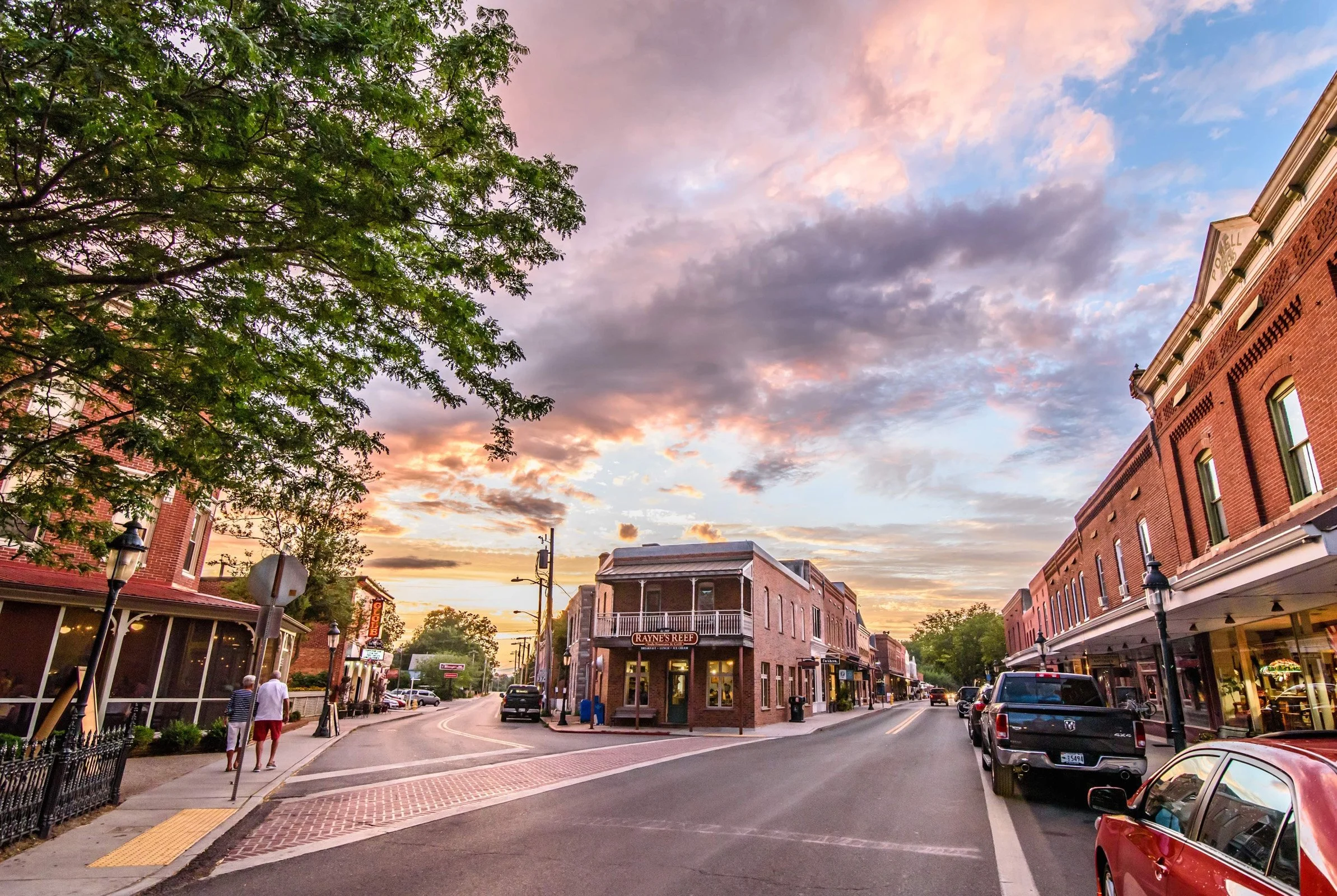 A small downtown street at sunset with brick buildings, parked cars, and a few pedestrians. The sky is partly cloudy with pink and purple hues.