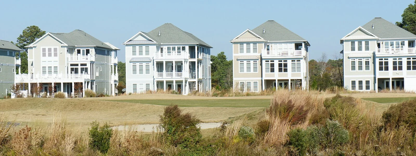 A row of five modern multi-story houses with white and pastel exterior colors, set on a grassy landscape under a clear blue sky.