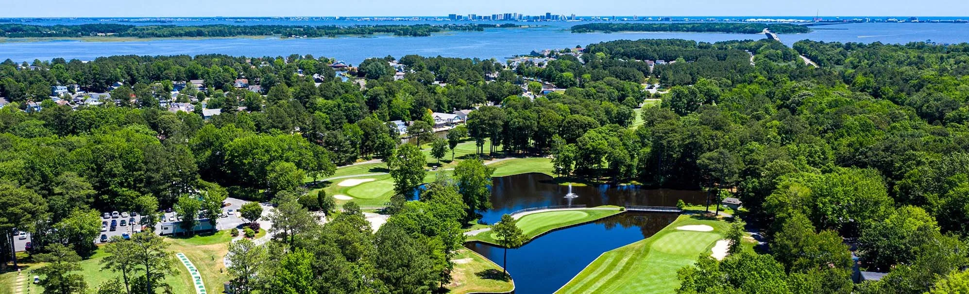 Aerial view of a golf course surrounded by dense trees, with a pond and fountain in the center, and a city skyline across a large body of water in the background.
