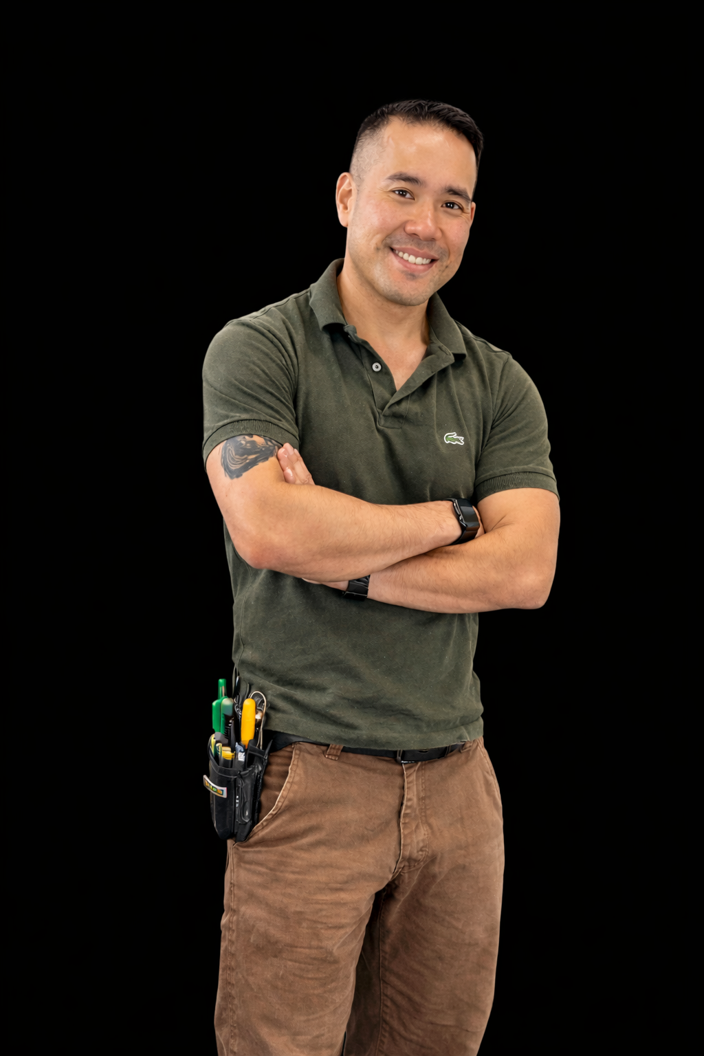 A man standing with arms crossed, smiling, wearing a dark green polo shirt with a small crocodile logo, brown pants, and a tool belt with various tools, against a black background.