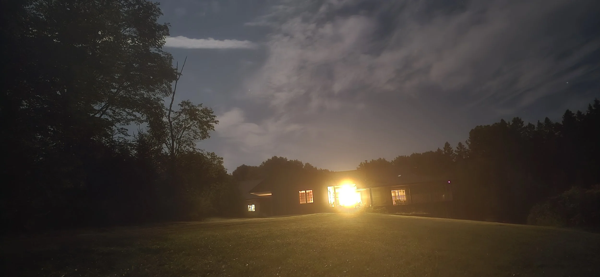 Nighttime scene of a house with lights on, surrounded by trees with a cloudy sky overhead.