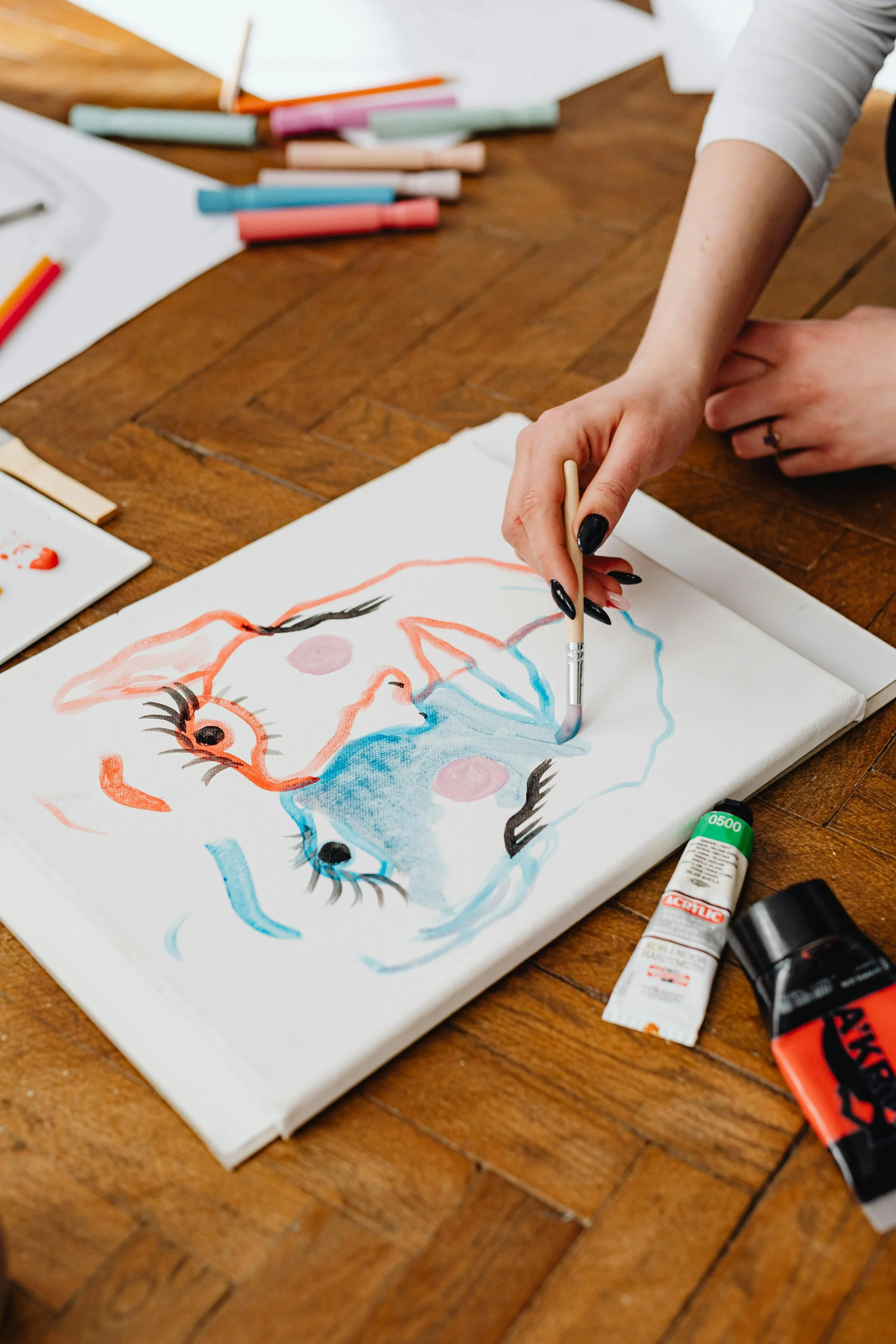 Person painting a colorful abstract face on canvas with various art supplies on a wooden table.