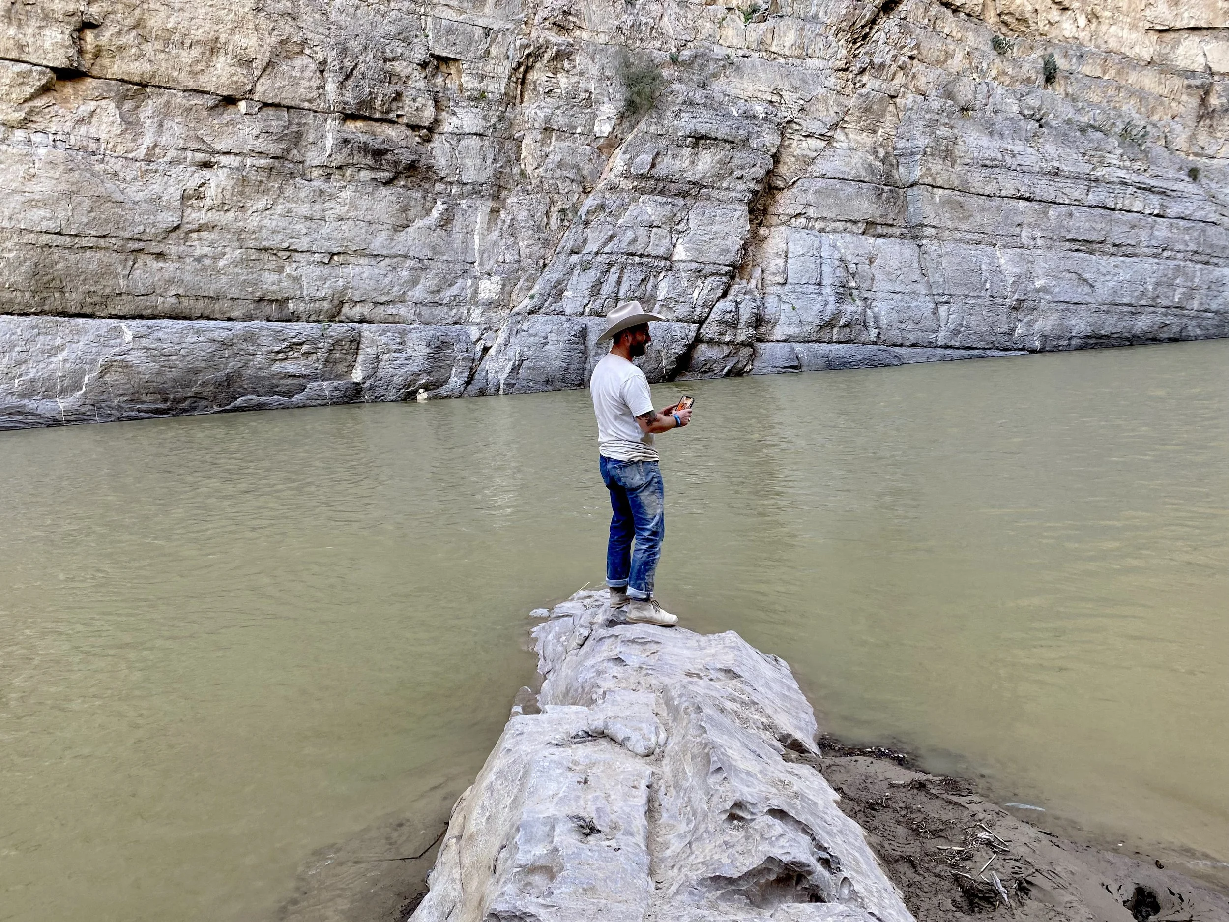 A man wearing a cowboy hat, white t-shirt, and jeans stands on a large rock extending into a body of water, holding and looking at a phone. Behind him is a rocky cliff wall.