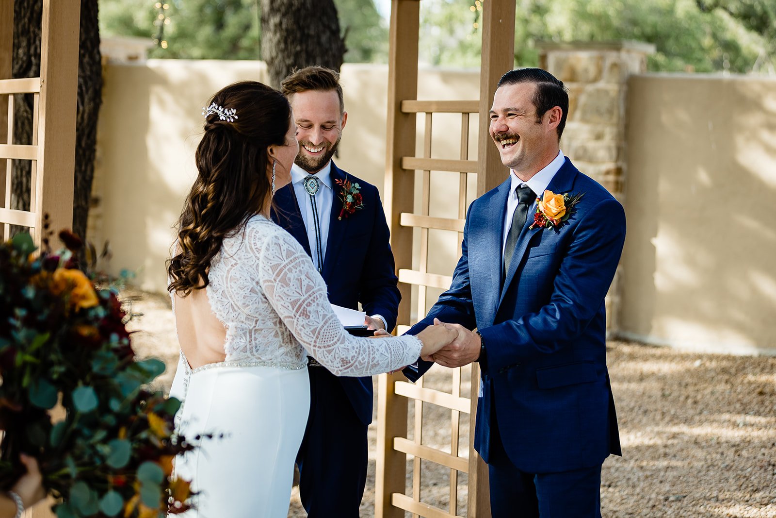 A couple getting married and holding hands at an outdoor wedding ceremony, with an officiant standing behind them, all smiling and dressed formally.