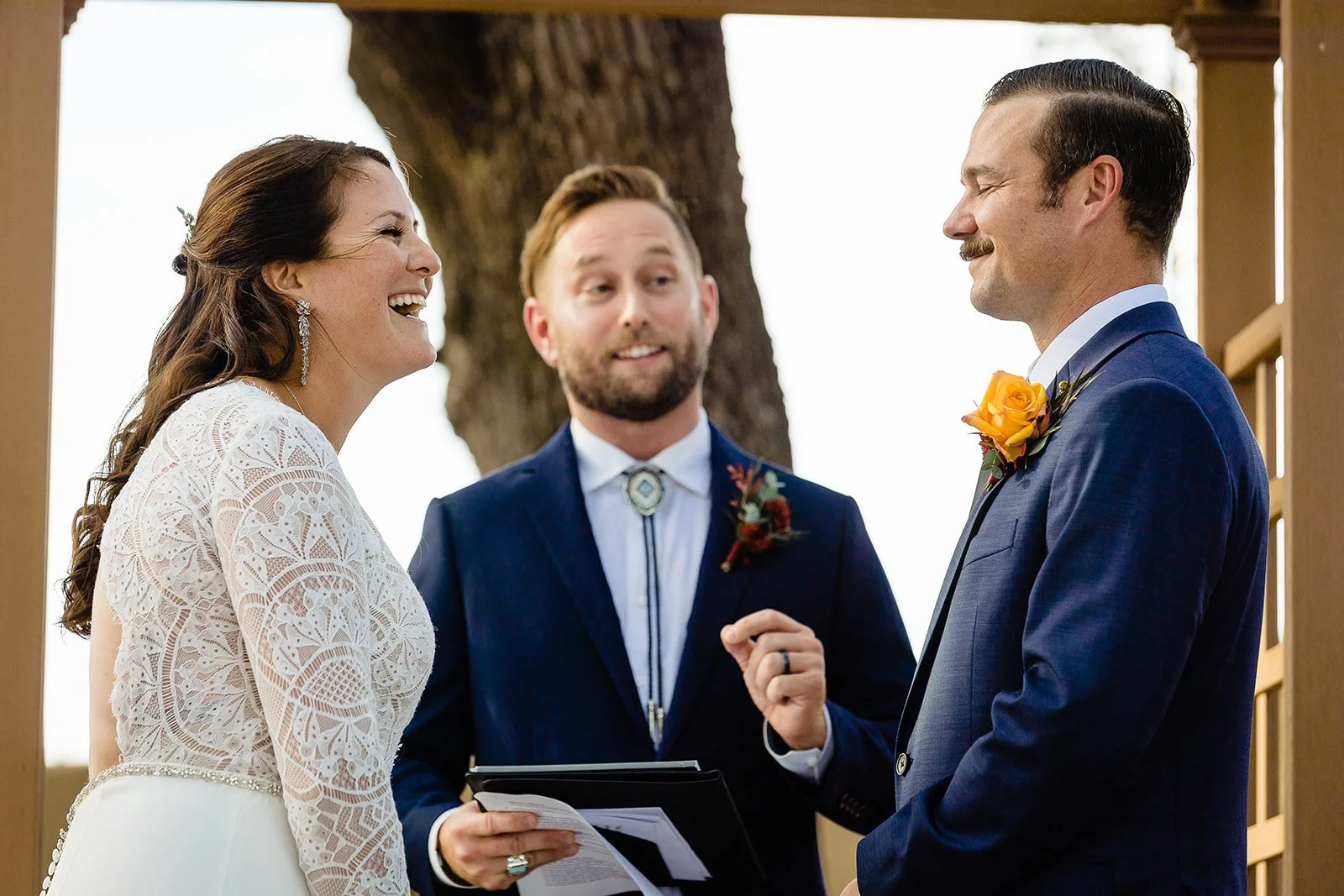 A smiling bride and groom exchanging vows during their wedding ceremony, officiant standing between them, outdoors with a large tree in the background.