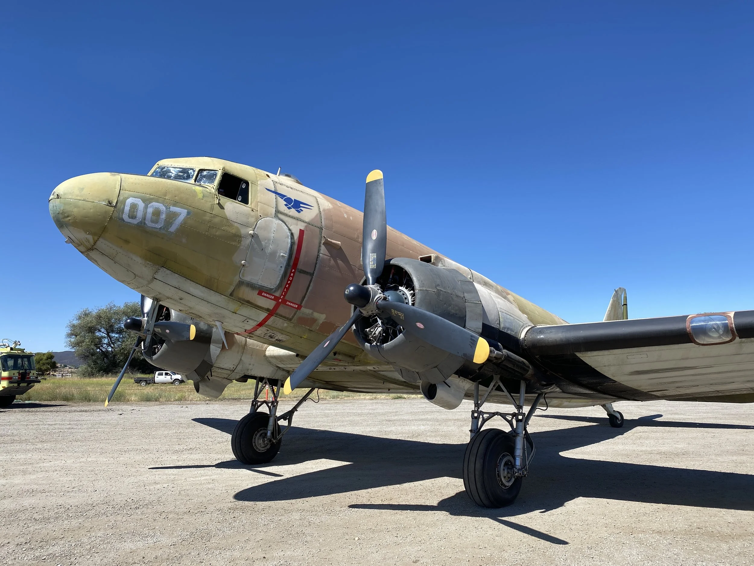 A vintage military aircraft with a camouflage paint job, parked on a gravel surface against a clear blue sky.