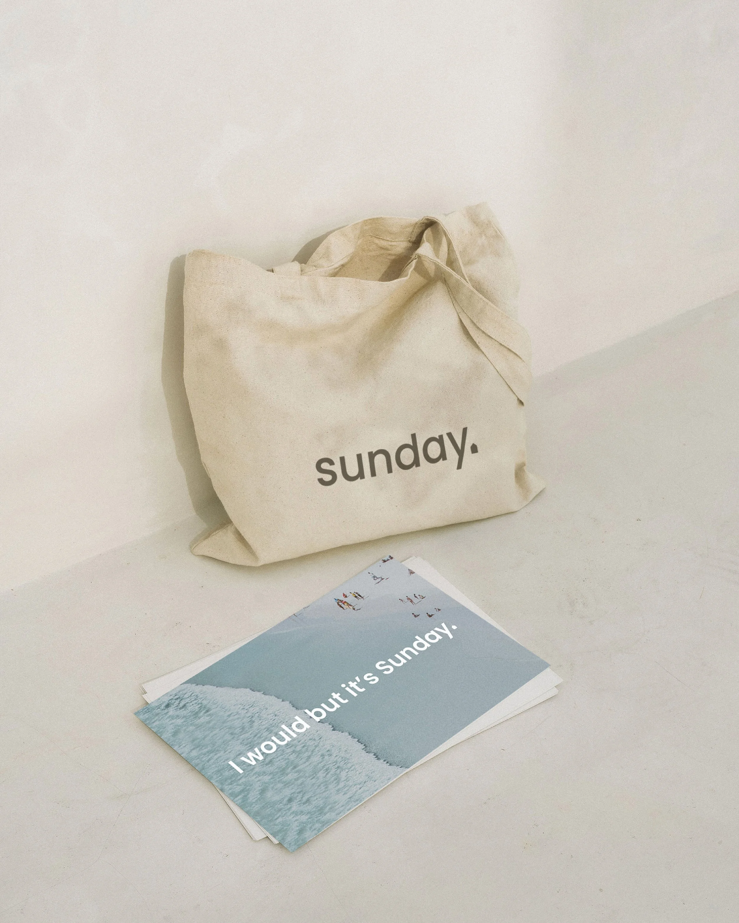 A beige tote bag with the word 'sunday.' printed on it, placed next to a paper with an image of a beach and the text 'I would but it’s Sunday.'