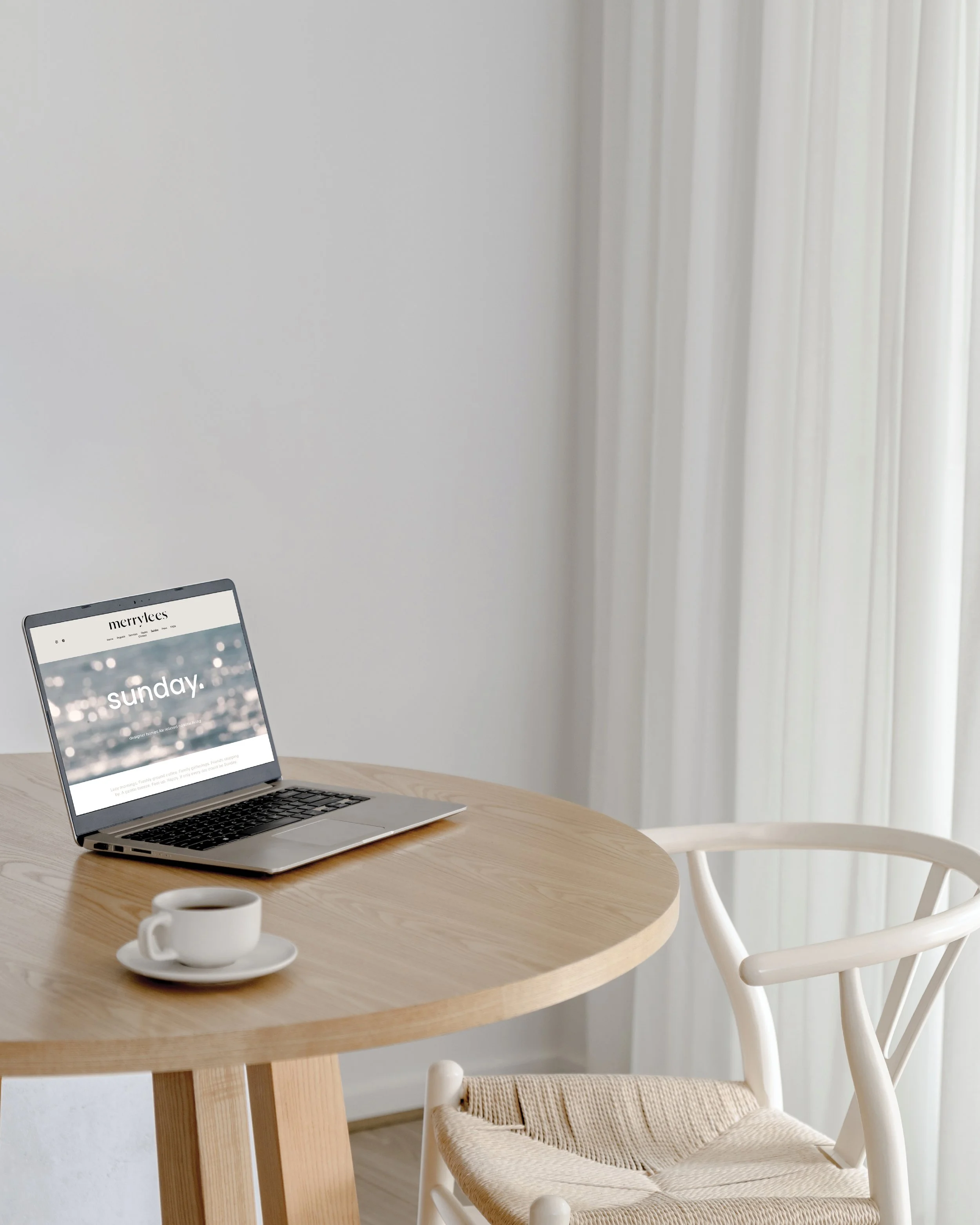 Minimalist workspace with a wooden round table, a laptop showing a website, a white coffee cup with saucer, a woven chair, and white curtains in the background.