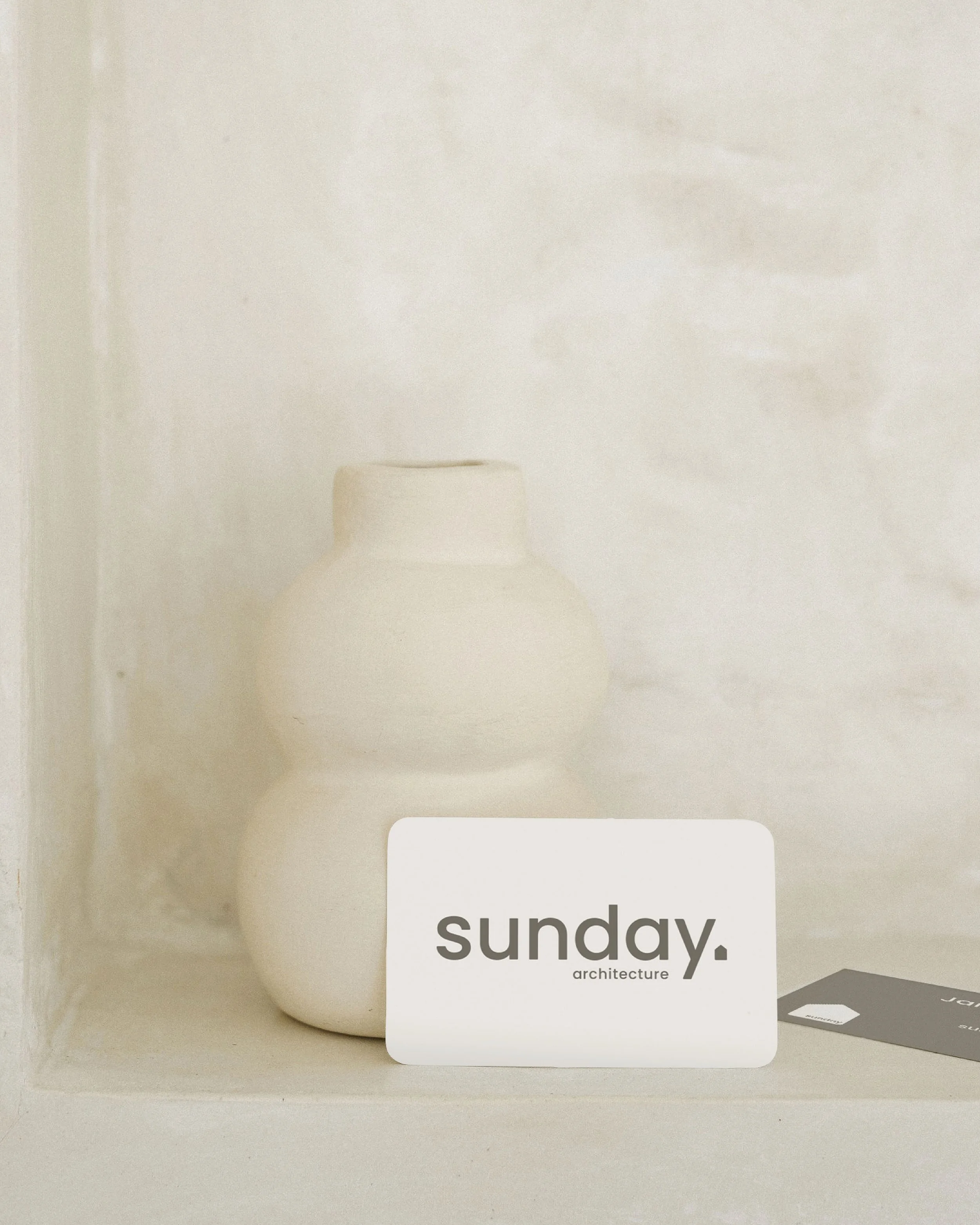 Minimalist white ceramic vase with a rounded design next to a business card with the text 'sunday. architecture' on a textured neutral background.