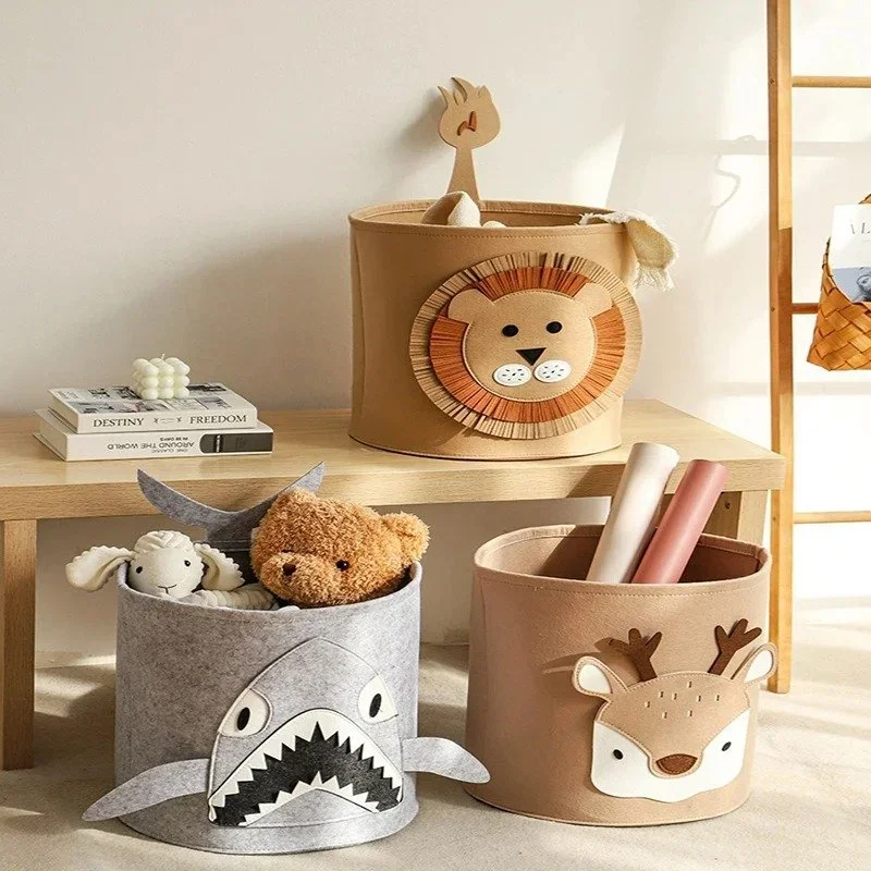 Three fabric storage baskets with animal designs on a wooden table and floor, filled with plush toys and paper rolls, with a small wooden rack and books in the background.