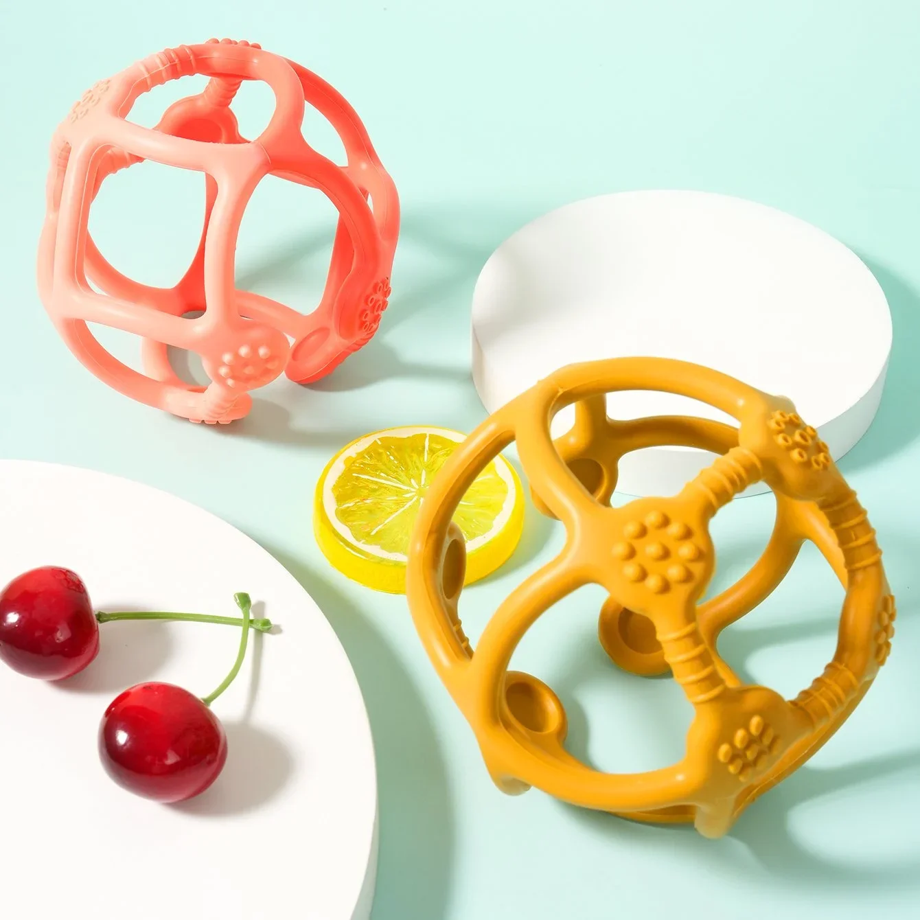 Colorful spherical baby teething toys, a slice of lemon, and cherries arranged on a pastel background with white platform surfaces.