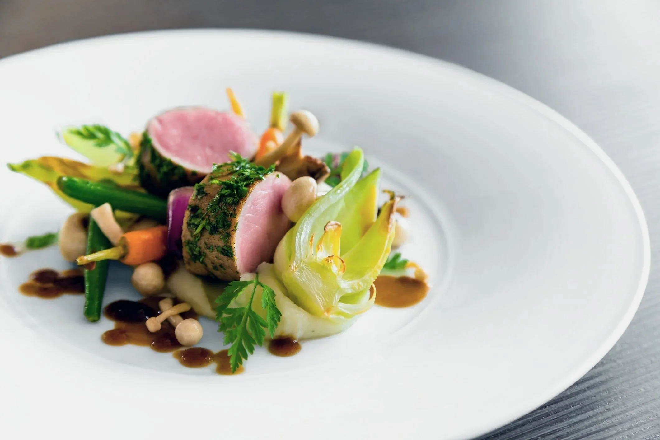 A gourmet dish featuring sliced seared meat, vegetables, herbs, and sauce served on a white plate.