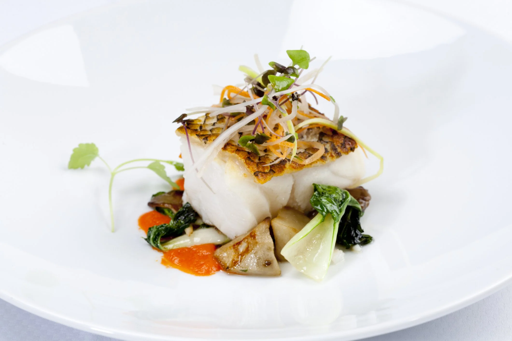 A gourmet seafood dish with a piece of white fish, caviar, microgreens, and roasted vegetables on a white plate.